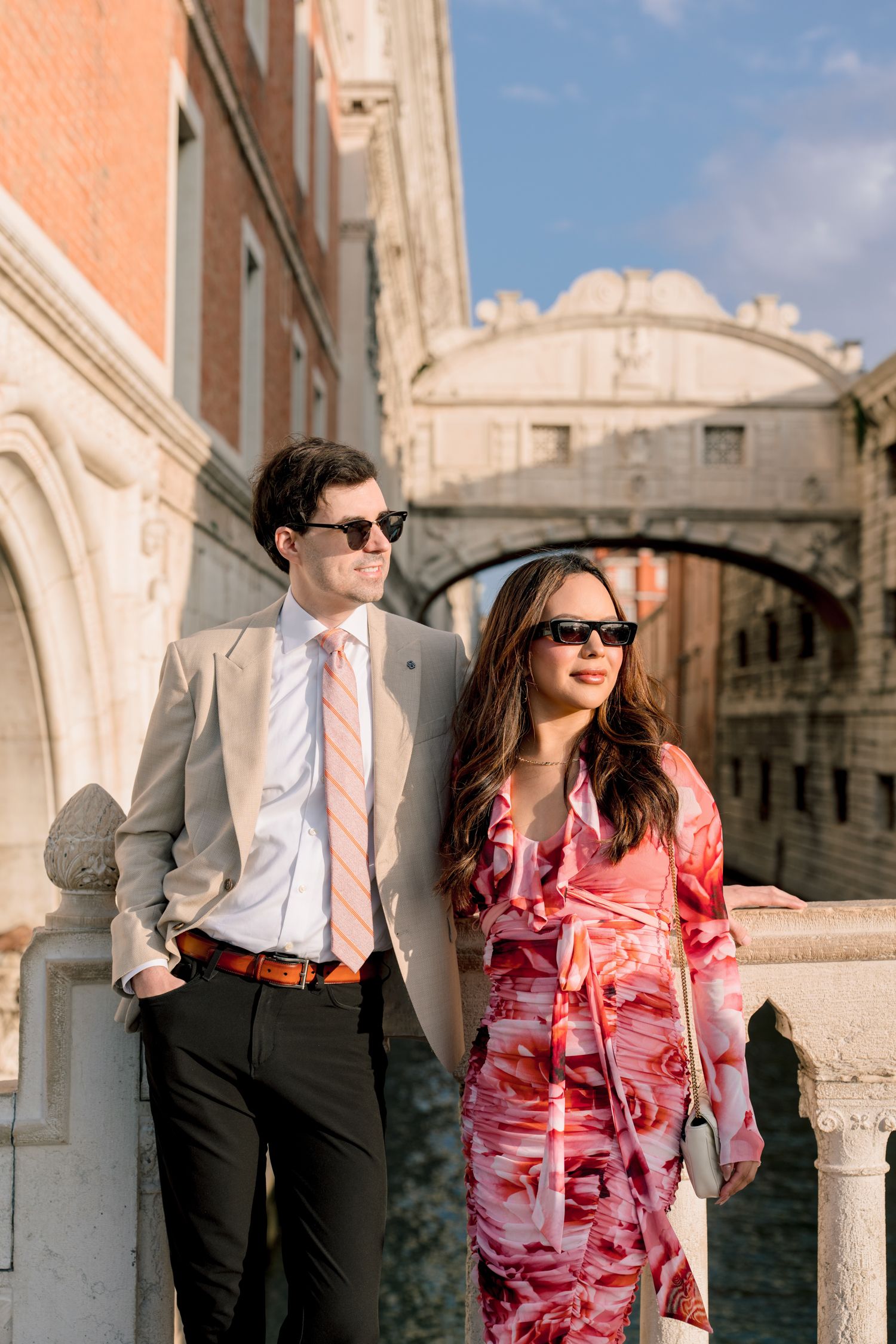 A stylish couple in formal attire poses along a historic stone balcony during a photoshoot in Venice.