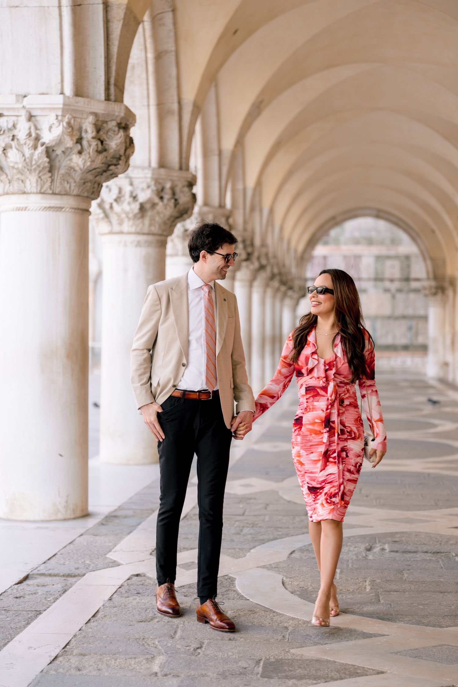 A couple walks through an elegant arched colonnade hallway in Venice, with the person in a coral dress twirling playfully.