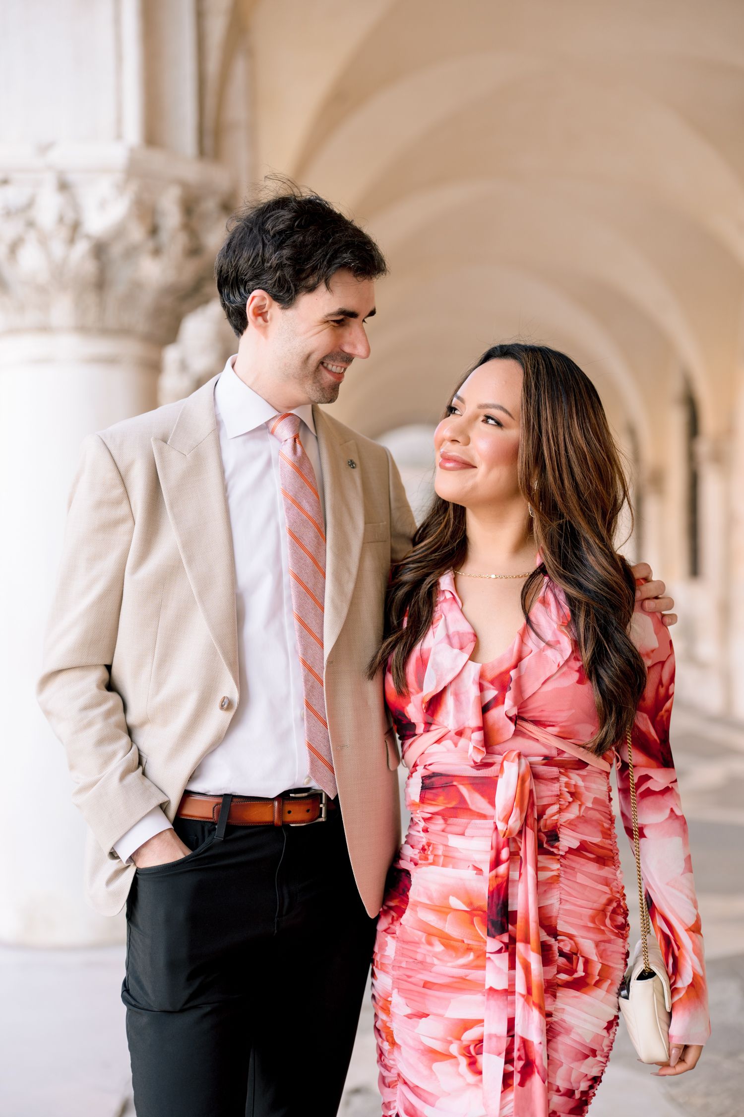 A couple in coordinating pink and beige outfits share a romantic moment under an ornate archway in Venice.