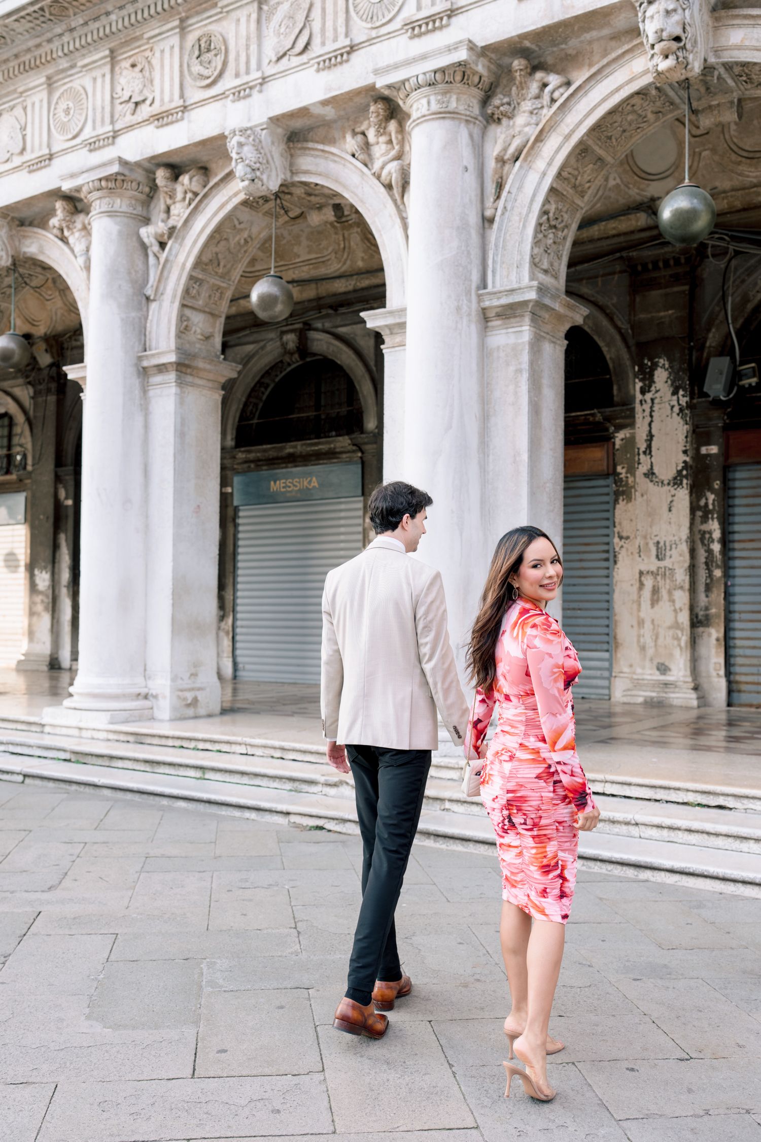 A couple walks through an ornate colonnade with classical architecture and stone arches in Venice.