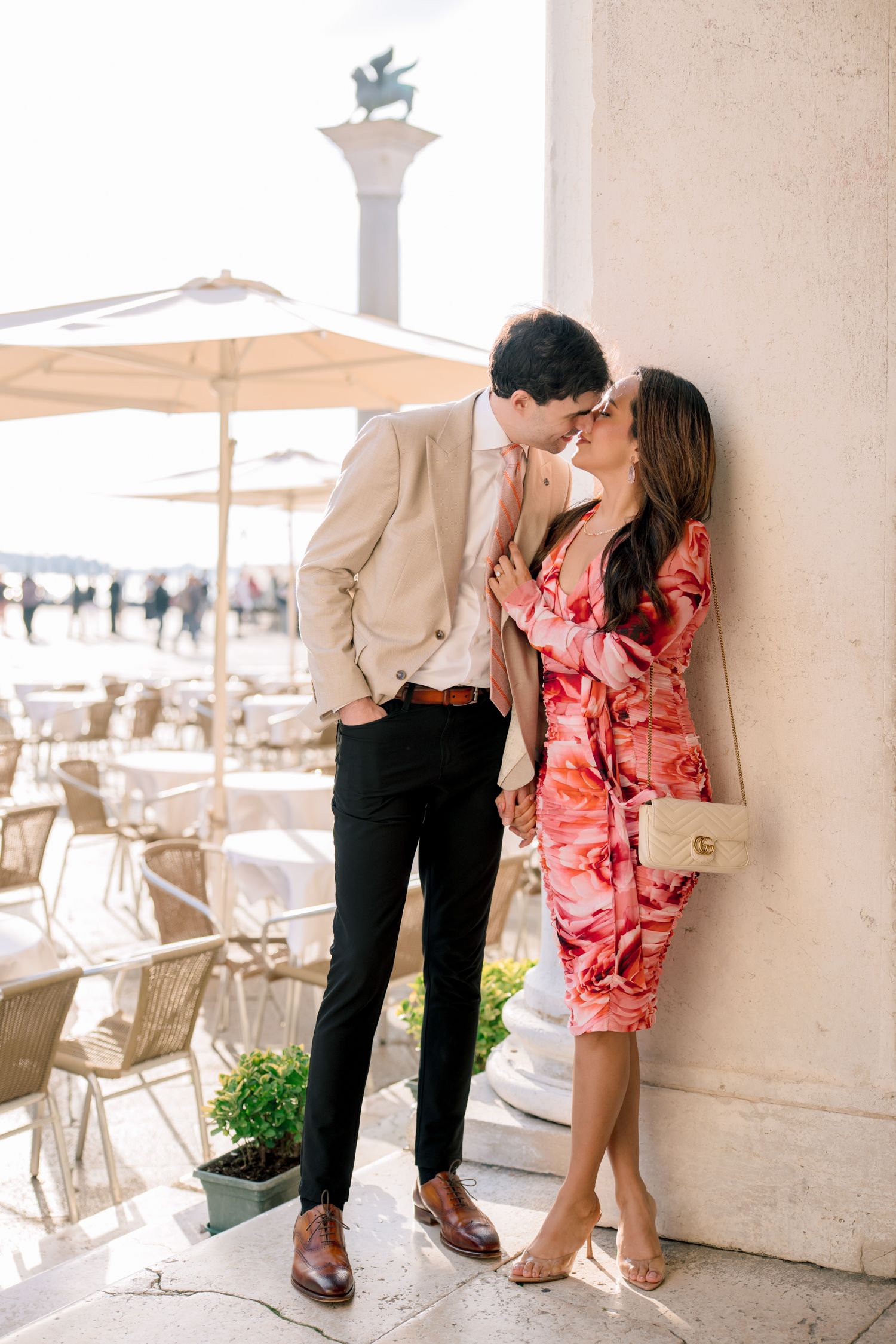 A romantic couple shares an intimate moment near St. Mark's Square in Venice with cafe seating visible.