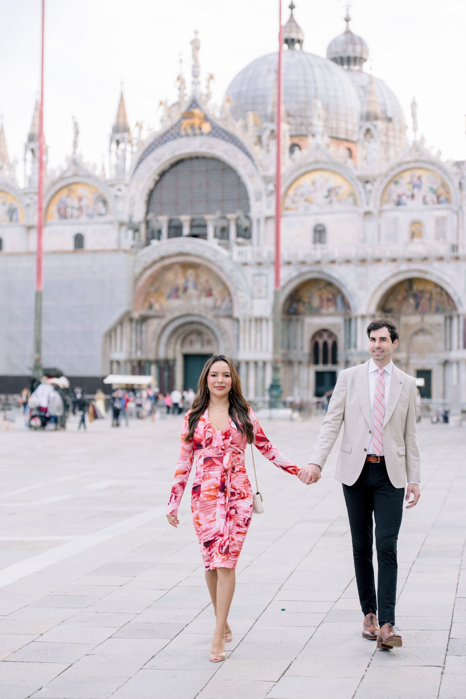 A couple holding hands walks in front of the stunning St. Mark's Basilica in Venice's famous piazza.