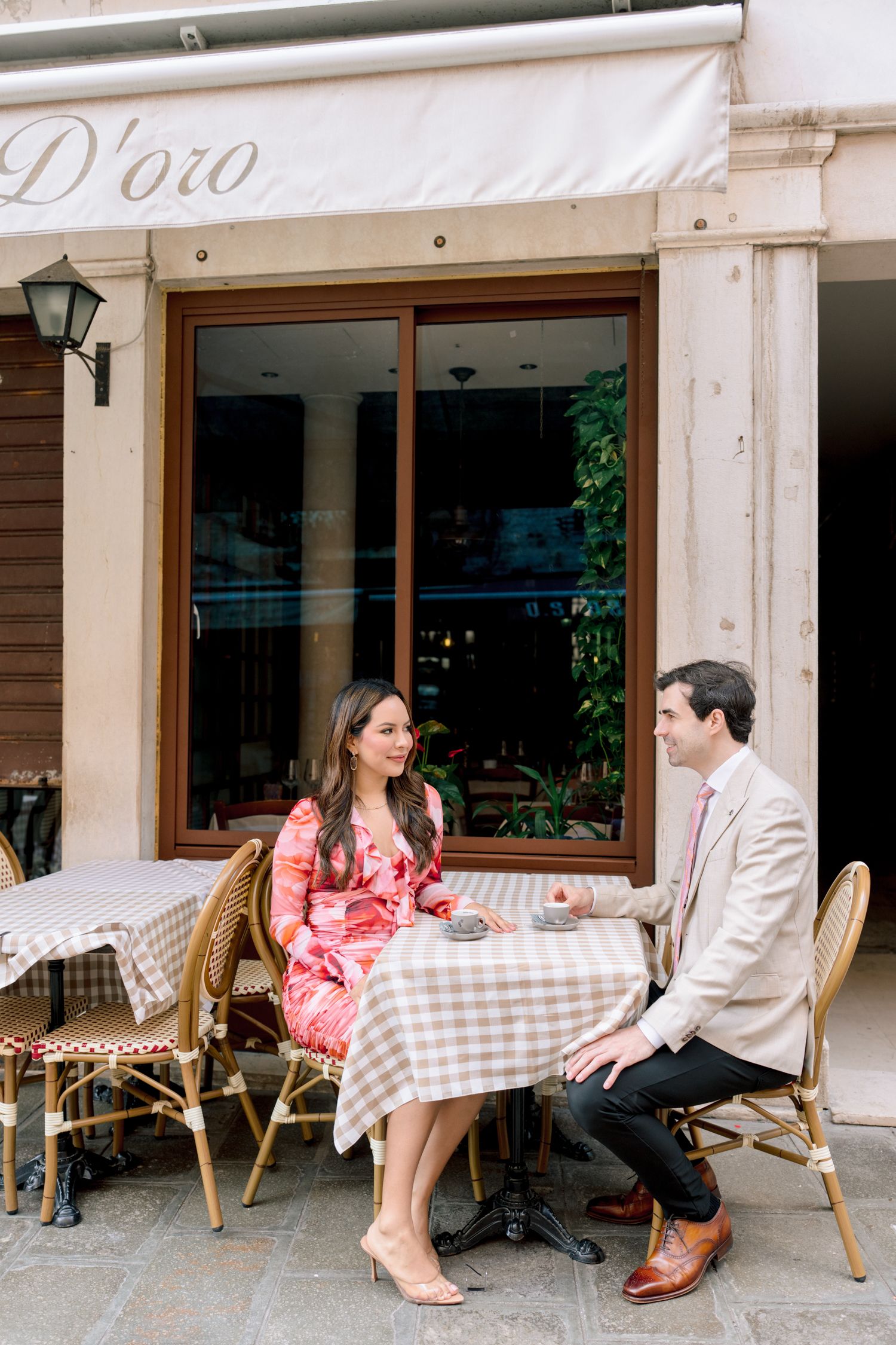 A couple sits at a cafe table with checkered tablecloth in a romantic outdoor setting.