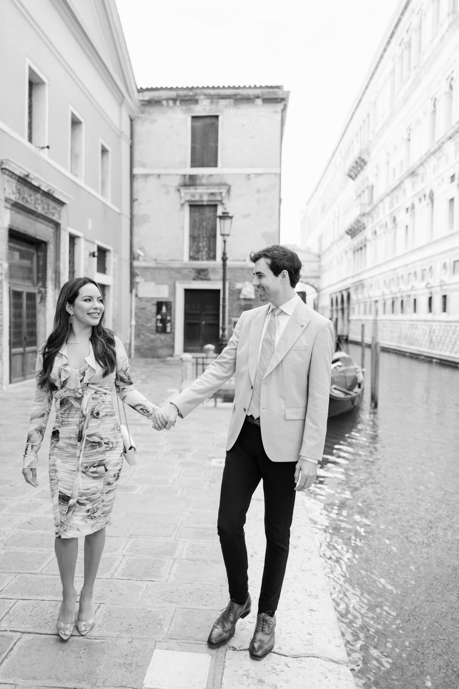 A romantic black and white photo of a couple walking along a Venice canal, with classic Italian architecture in view.