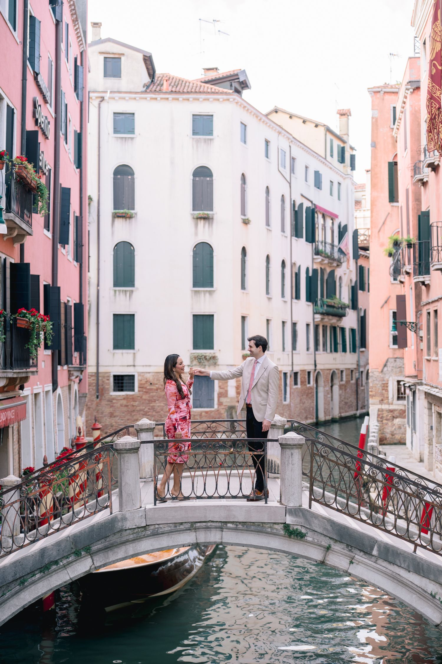 Two people standing on a romantic bridge over a canal in Venice with pink and white buildings in the background.