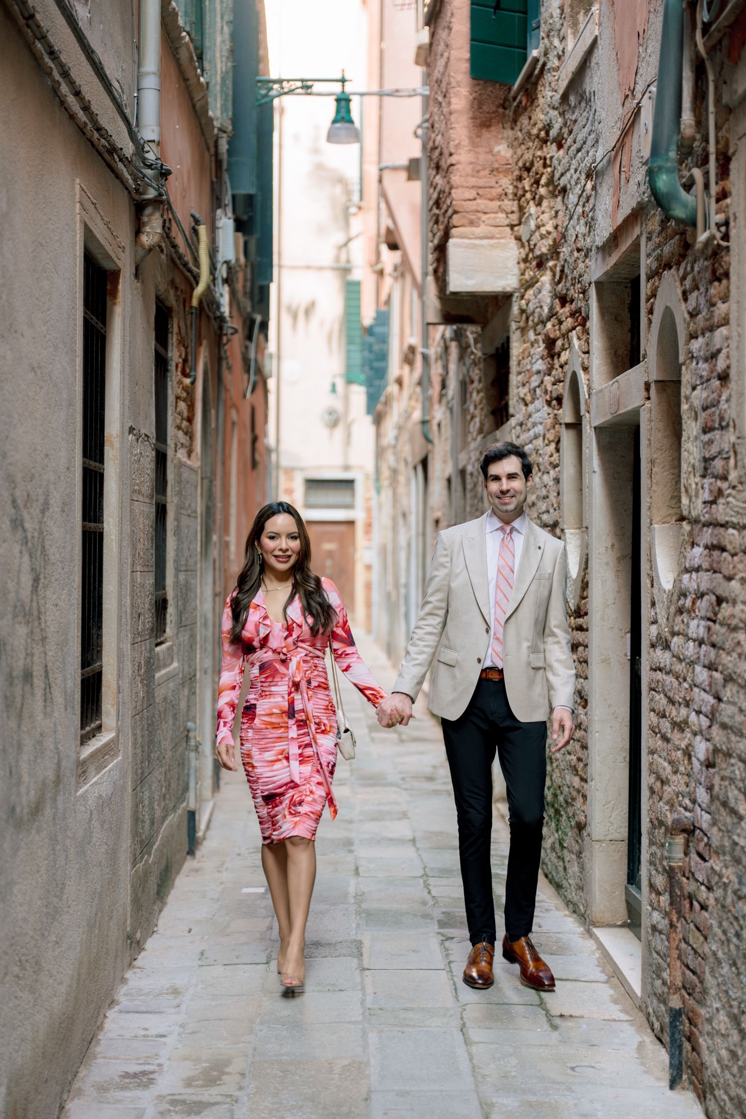 A couple walks hand in hand through a narrow alley in Venice with stone walls and green shutters.