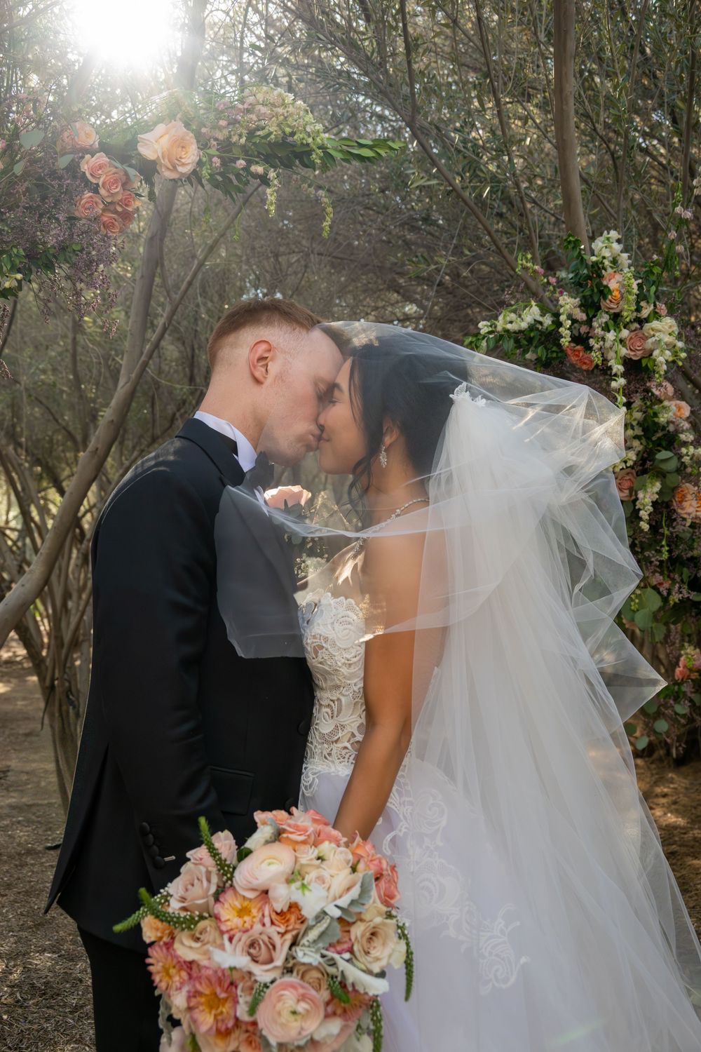 Bride and groom share first kiss under floral arch with veil draped over them in forest wedding.