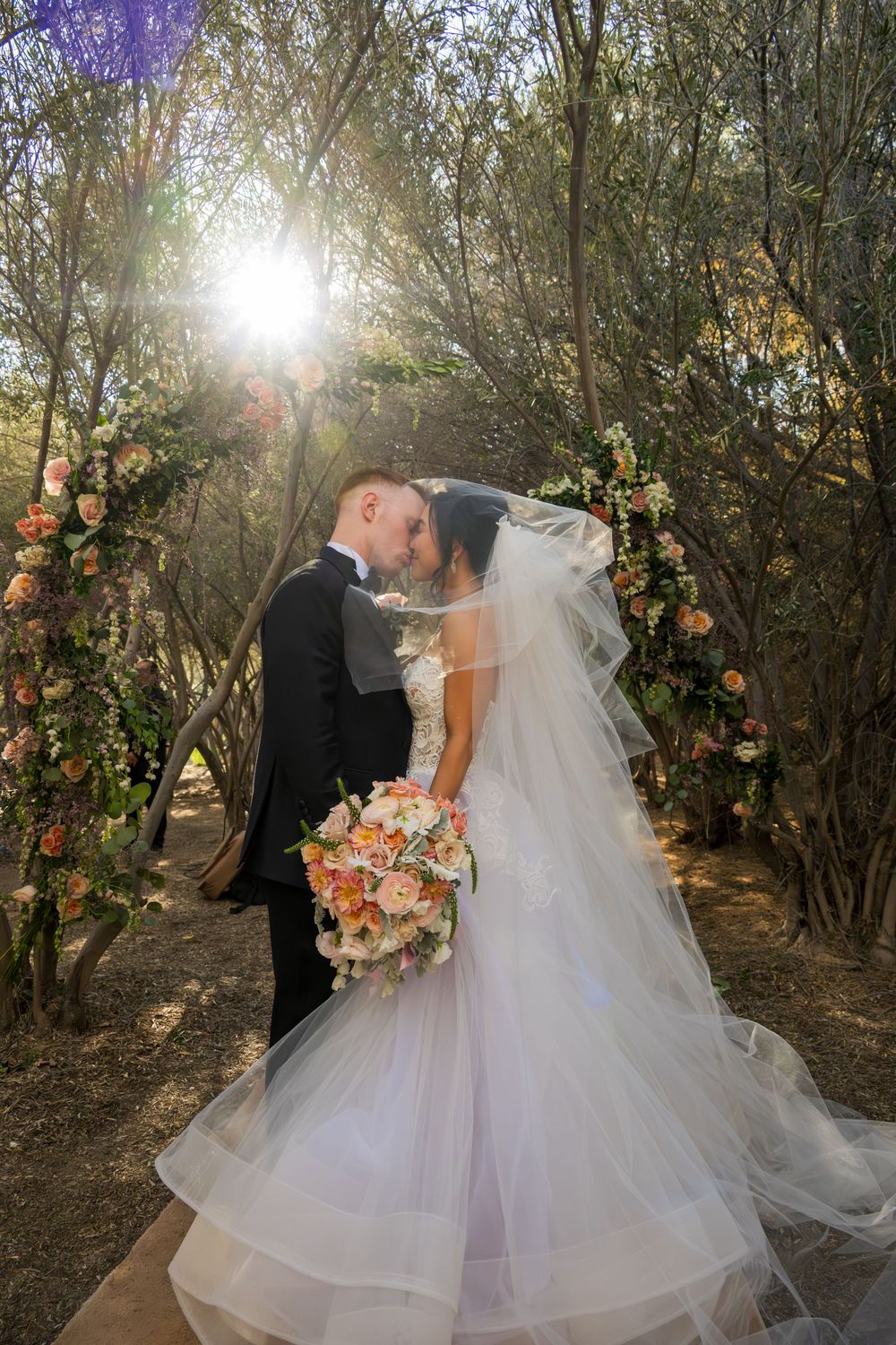 Newlyweds kiss beneath sunlit floral arch in woodland wedding ceremony with cascading bridal veil.