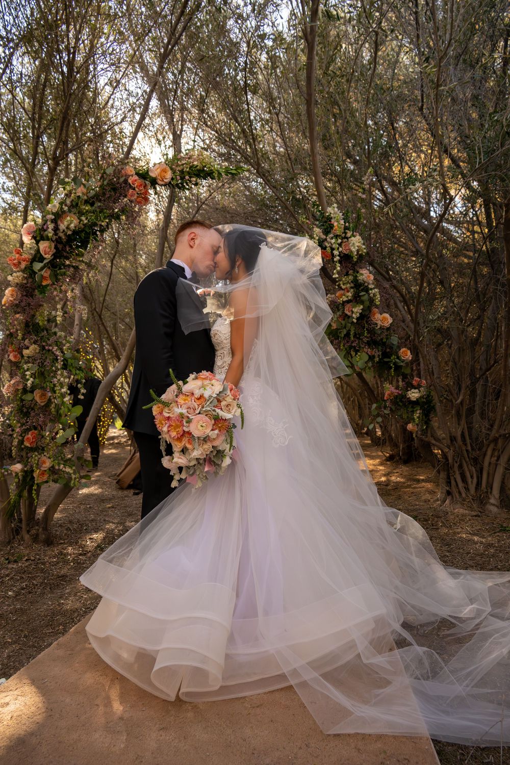 Bride and groom kiss at outdoor floral arch altar surrounded by trees in natural wedding setting.