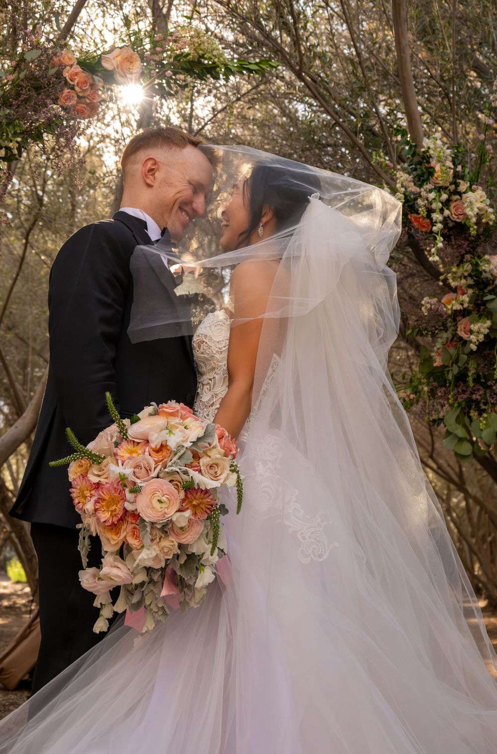 Smiling bride and groom touch foreheads tenderly beneath veil at woodland floral arch ceremony.