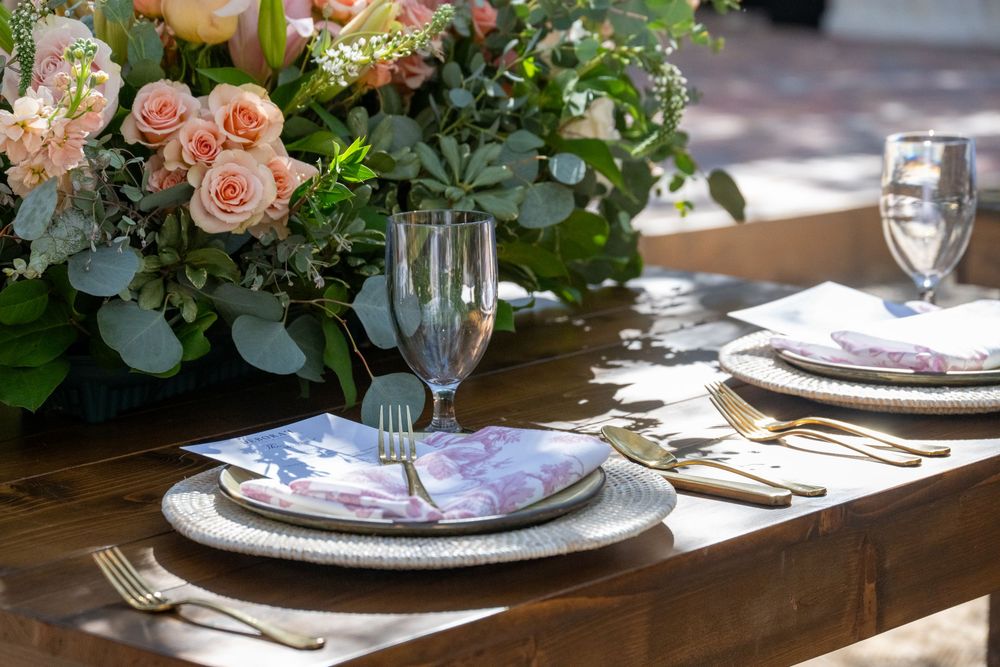 Elegant outdoor wedding reception table set with gold cutlery, floral napkins, and peach rose centerpiece.