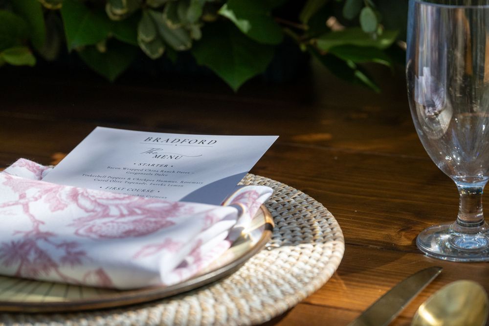 Close-up of a wedding menu card on a woven charger plate beside a crystal champagne flute.