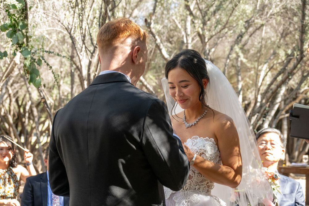Bride smiling as groom places wedding ring on her finger during outdoor woodland ceremony.