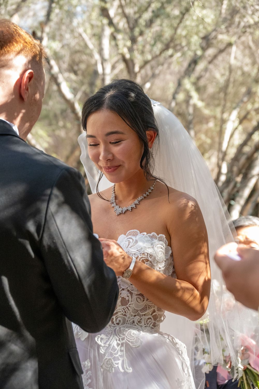 Radiant Asian bride smiling joyfully at groom during sunlit outdoor wedding ring exchange ceremony.