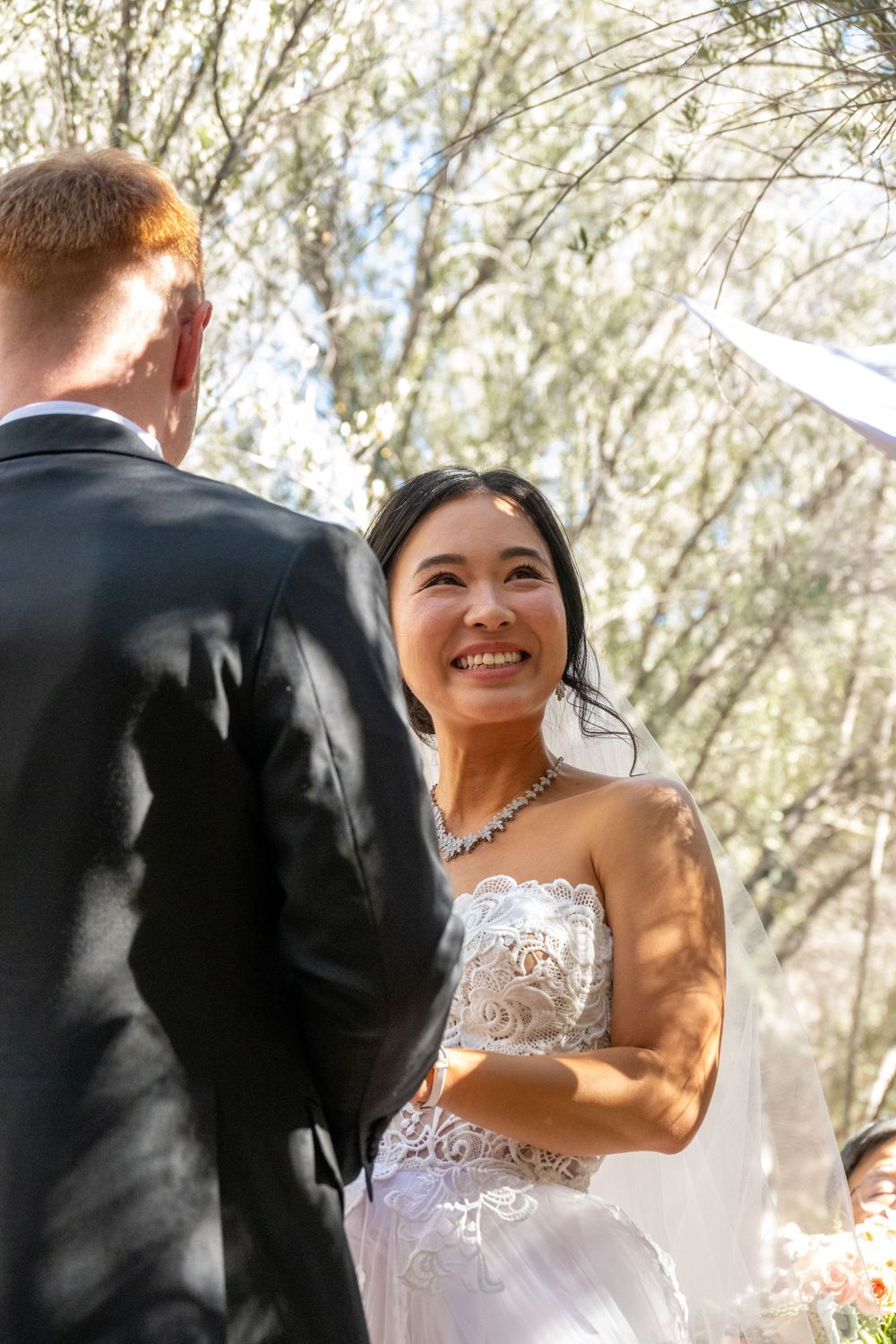 Smiling Asian bride gazing up at red-haired groom during sunny outdoor woodland wedding ceremony.