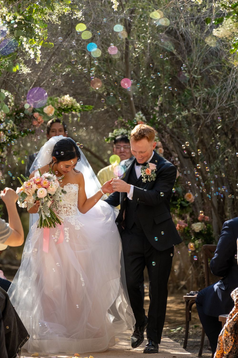 Bride and groom walk down outdoor aisle surrounded by colorful bubbles at woodland wedding ceremony.