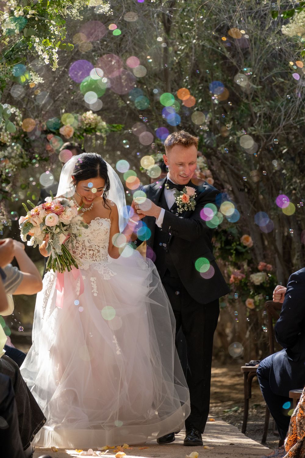 Newlyweds exit ceremony aisle through shower of colorful bubbles in sunlit forest wedding venue.
