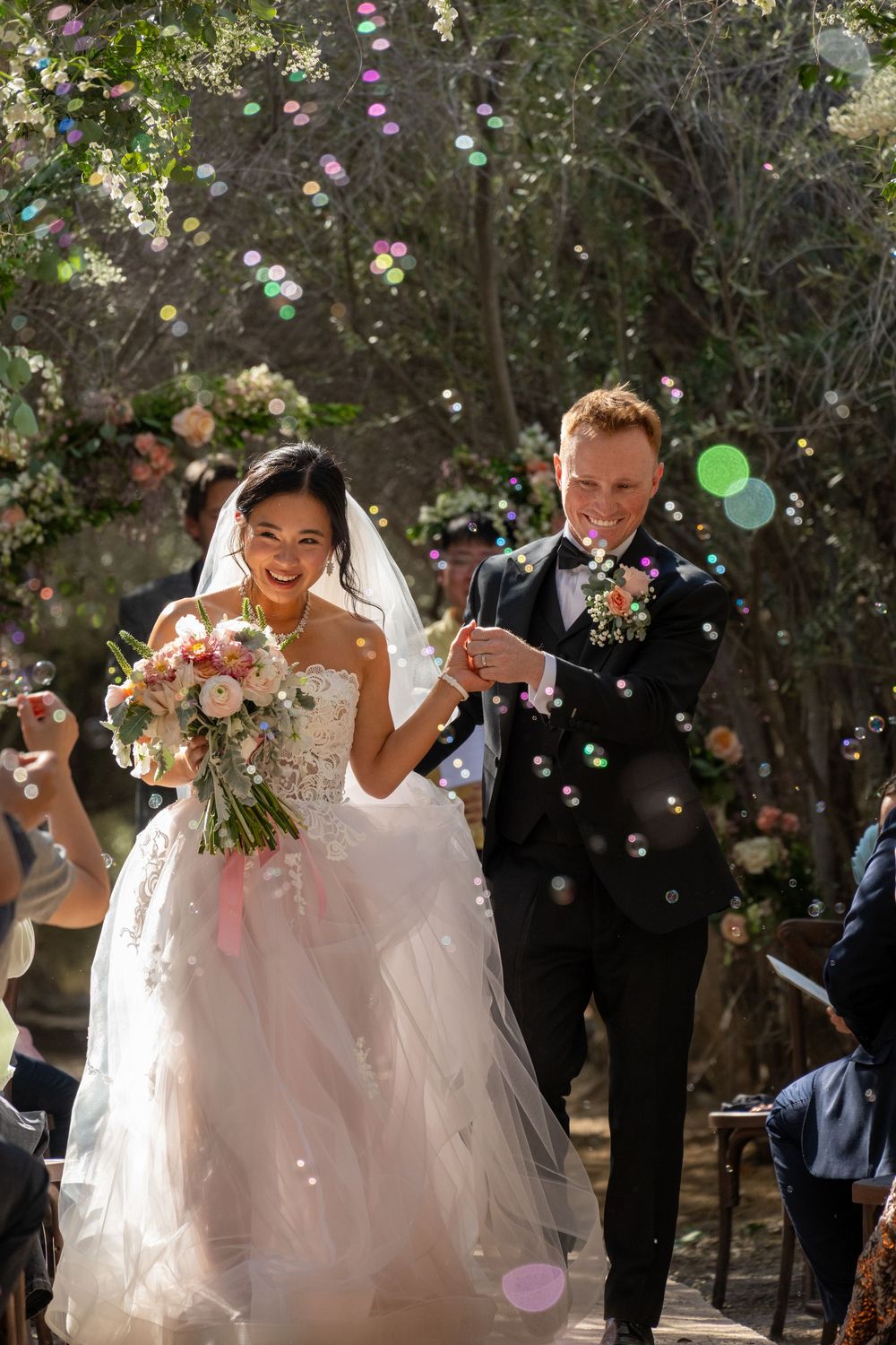 Smiling bride and groom walk hand in hand through bubbles at outdoor woodland wedding ceremony.
