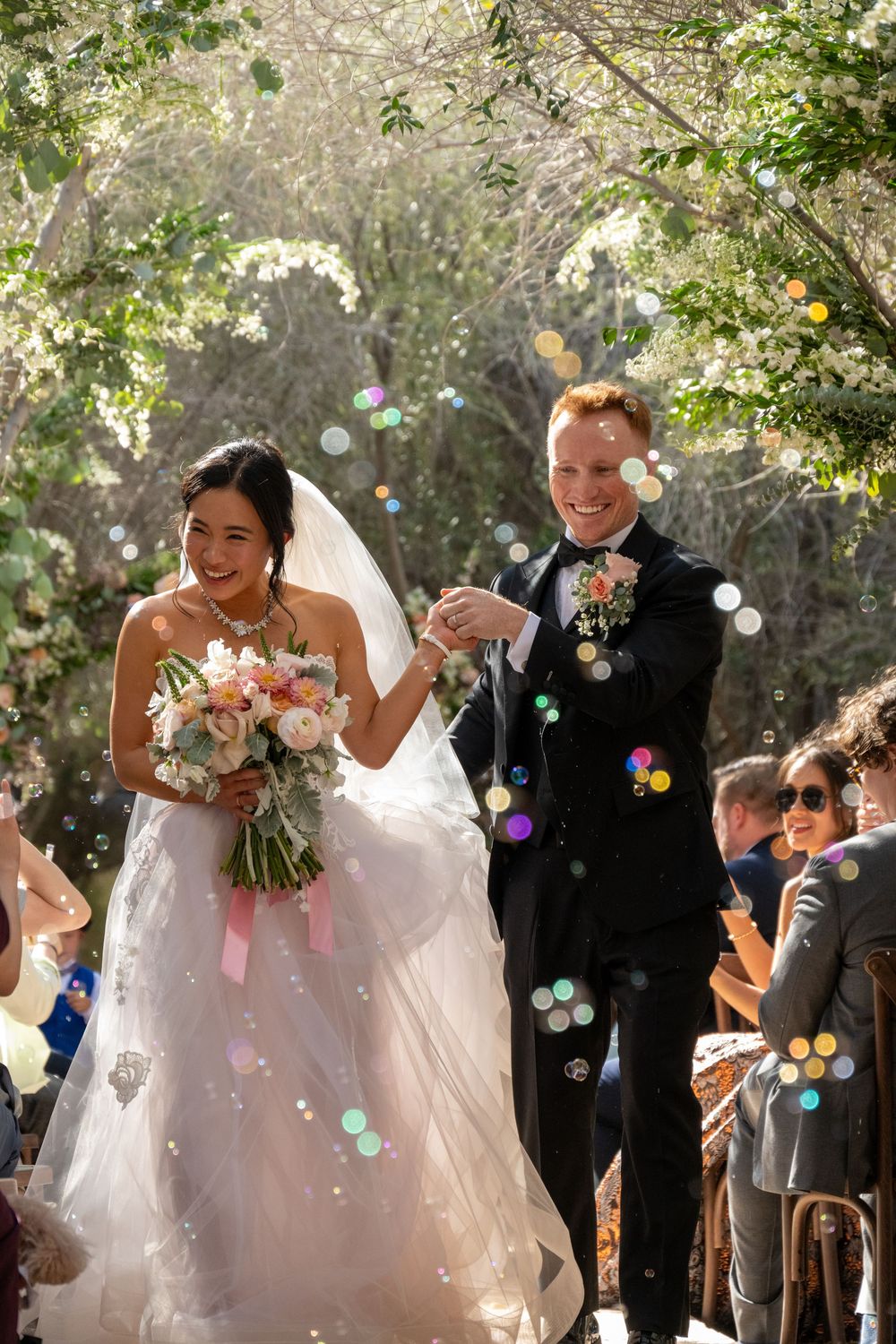 Joyful bride and groom exit wedding ceremony through bubbles in sunlit natural woodland setting.