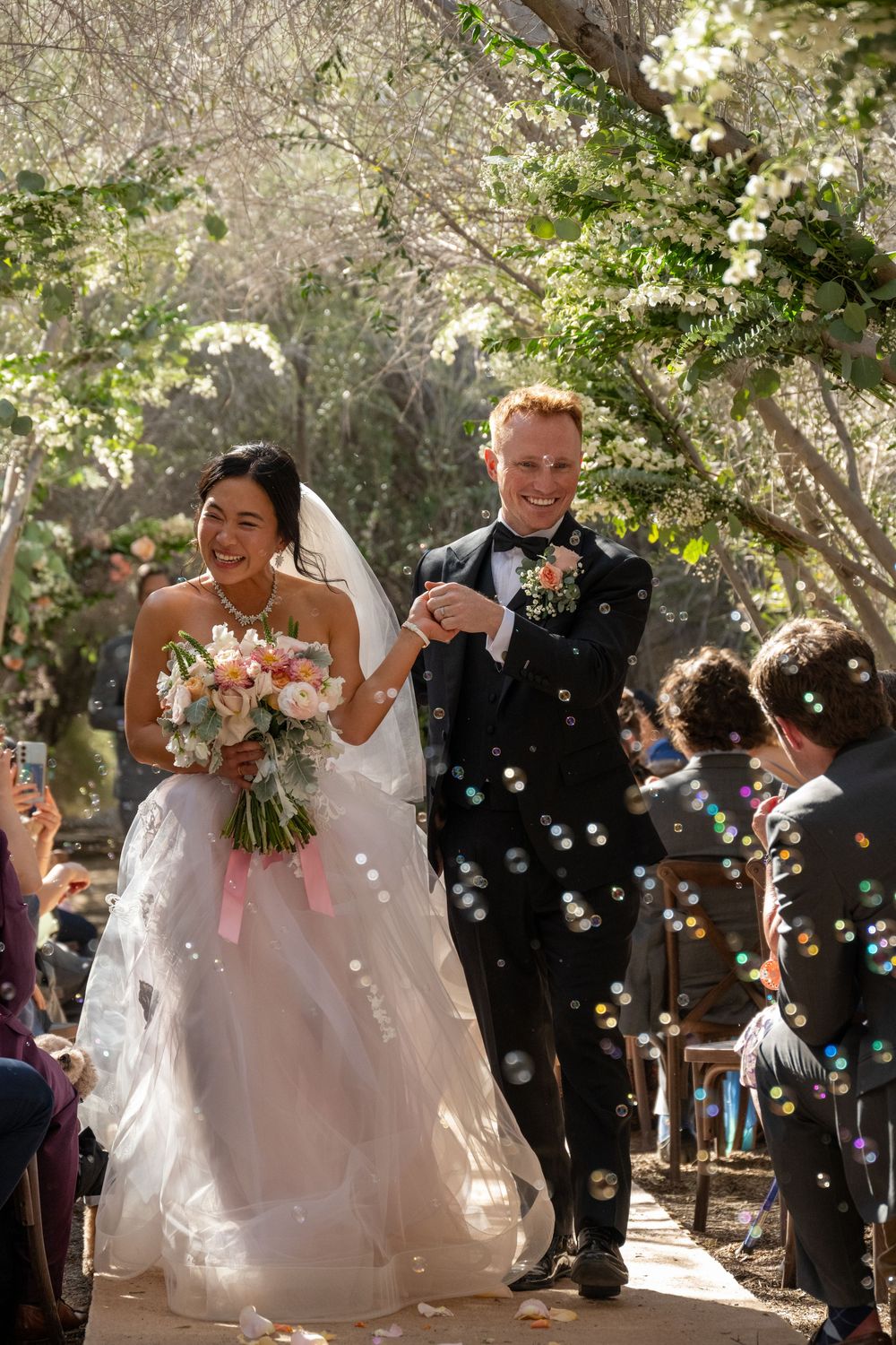 Happy newlyweds walk down tree-lined aisle with bubbles floating around them at outdoor wedding.