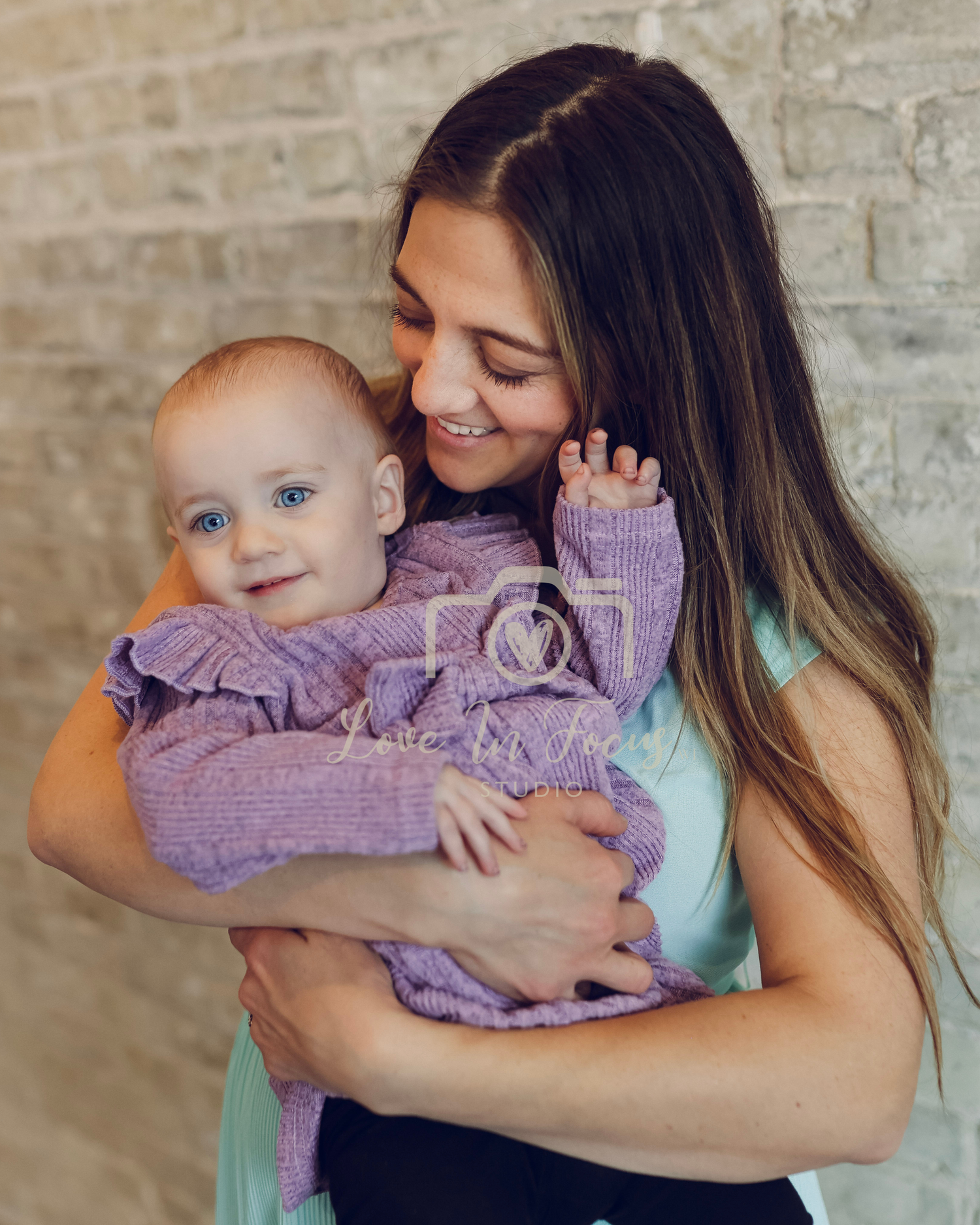 Young woman with long brown hair smiling while holding a happy baby dressed in a purple outfit.