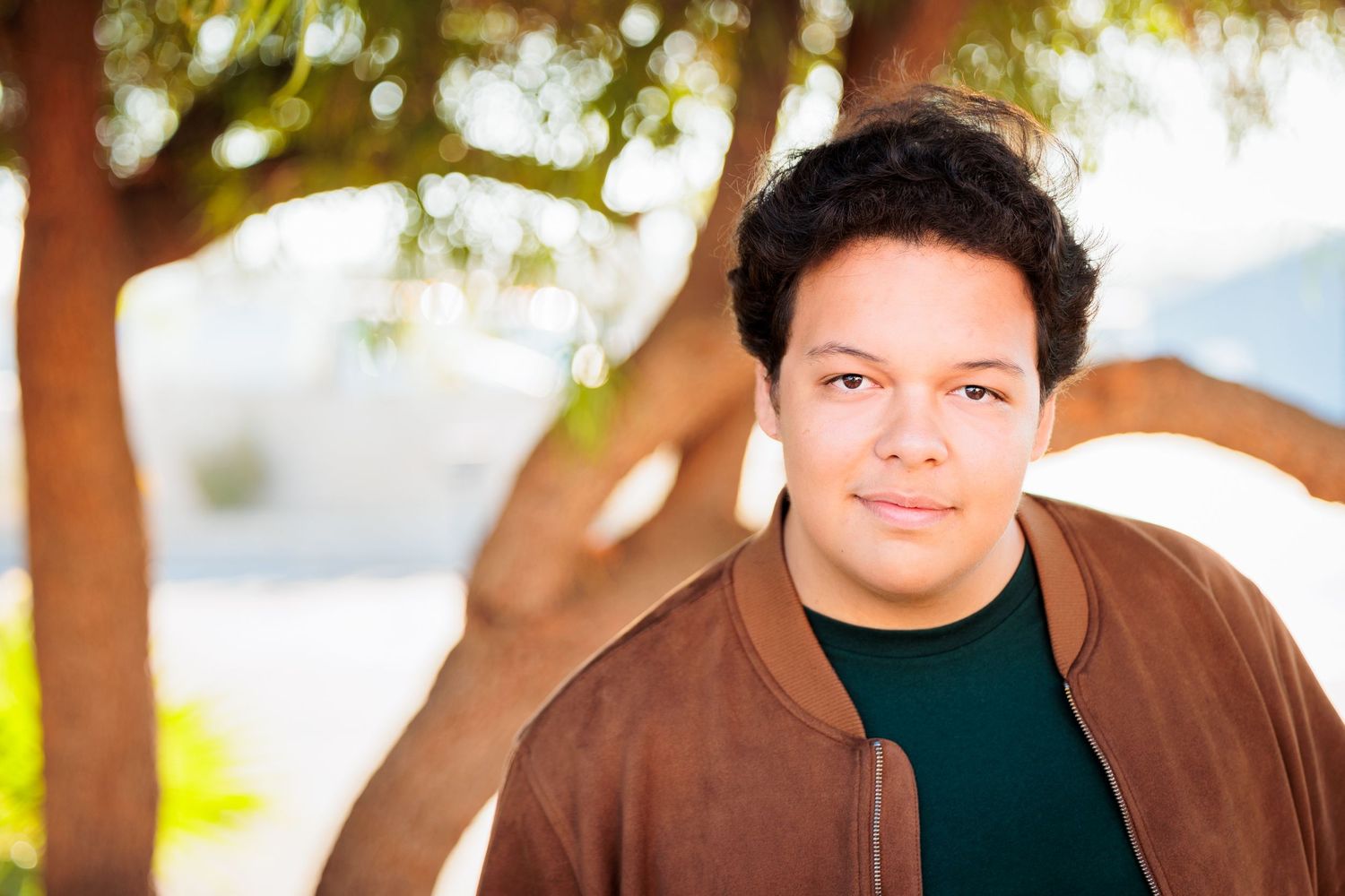 Teen boy in brown jacket poses outdoors under tree with warm bokeh background.