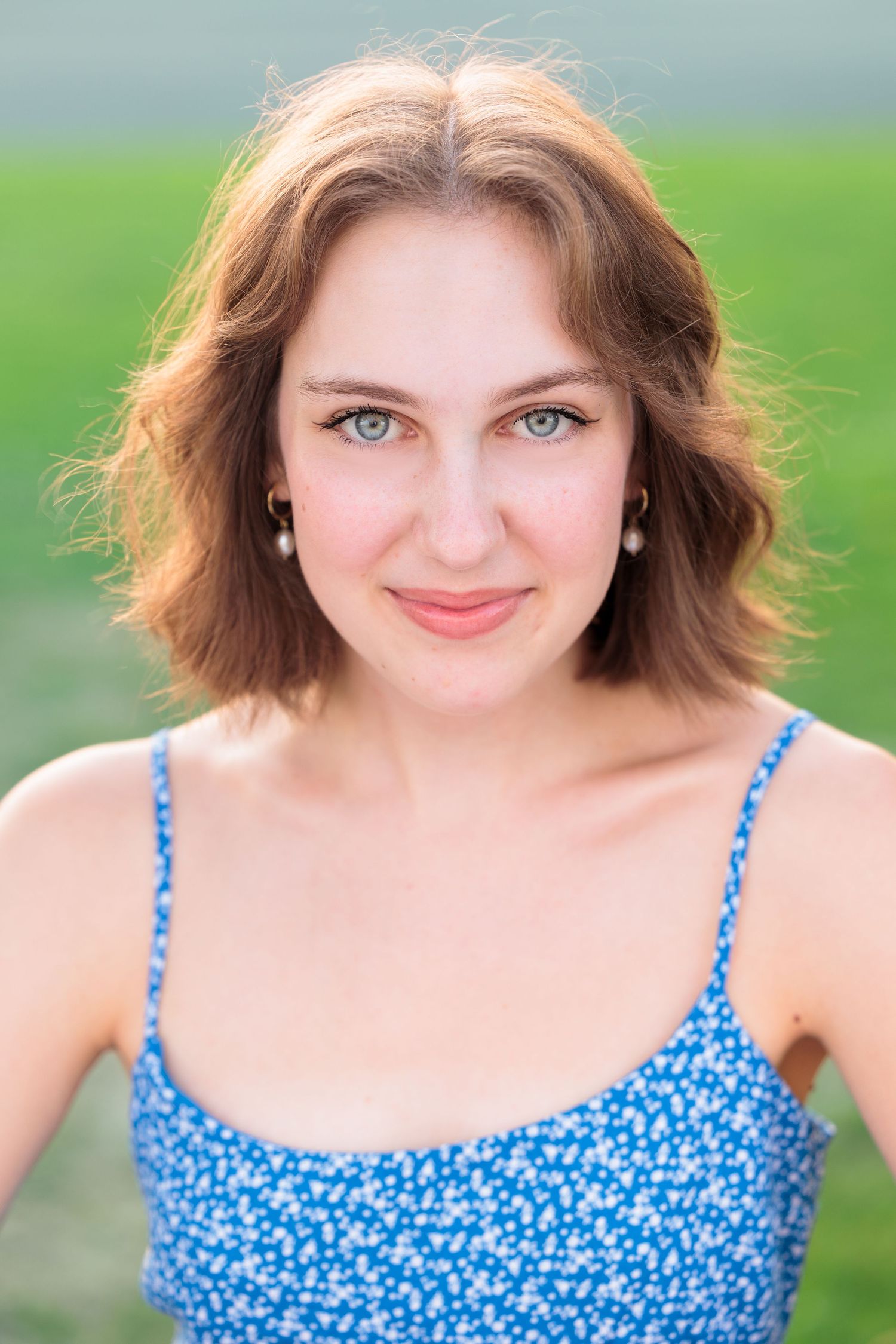 Young woman with blue eyes and auburn hair smiles in blue floral top outdoors.