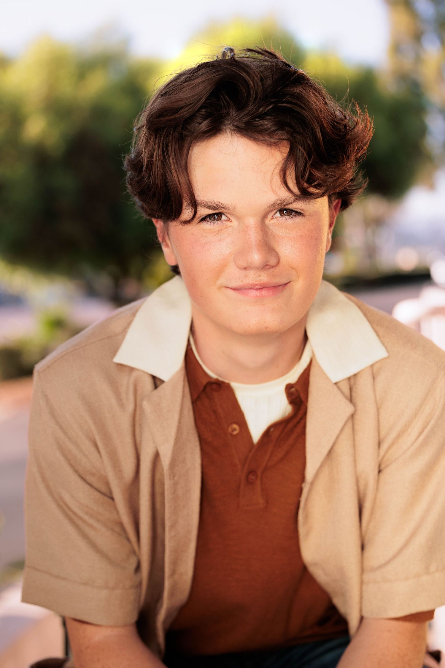 Teen boy with curly brown hair smiles in beige polo shirt outdoors in sunlight.