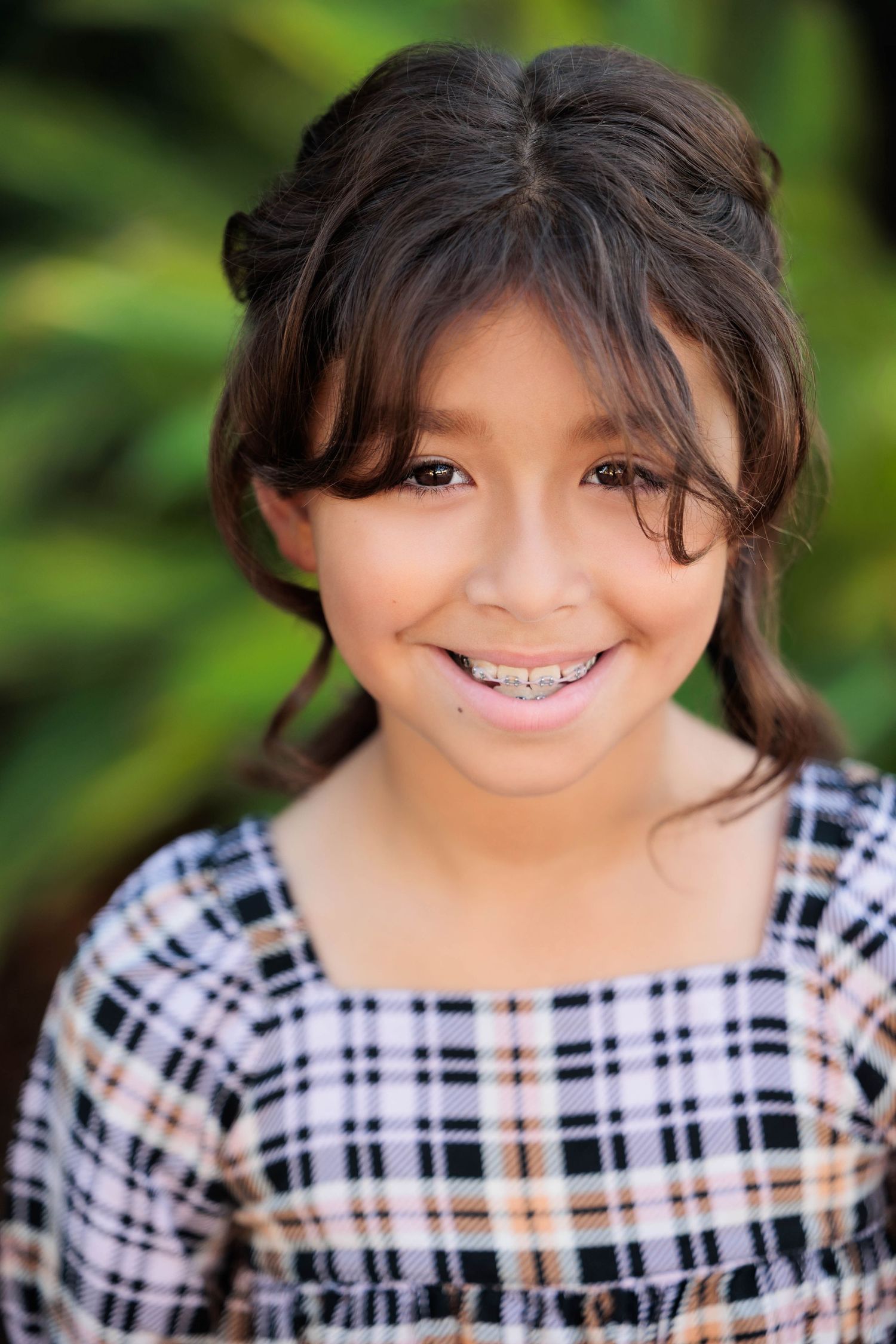 Smiling young girl with braces and dark hair wearing a plaid dress outdoors.