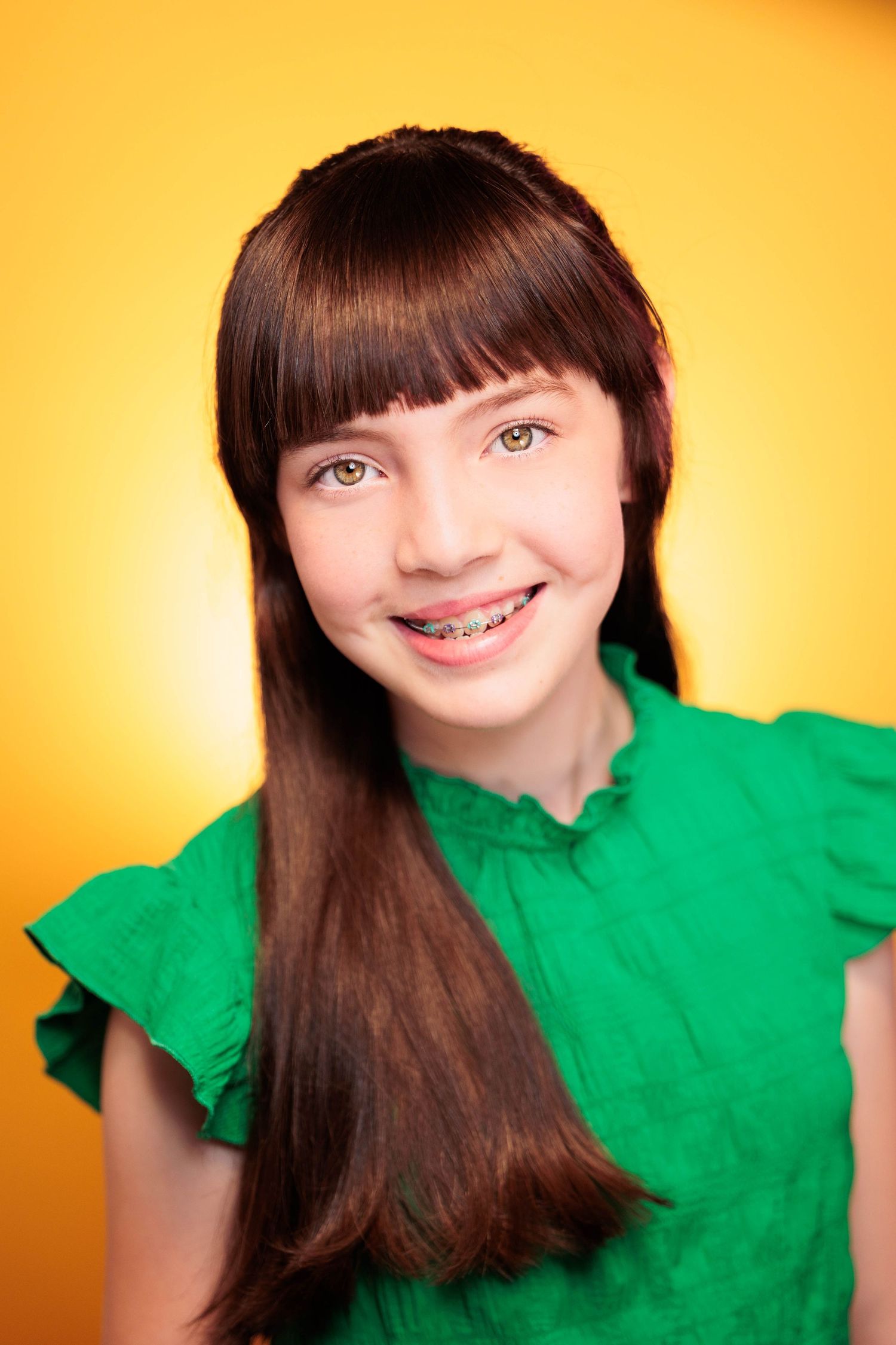 Smiling teenage girl with braces and long dark hair wears green top on yellow background.