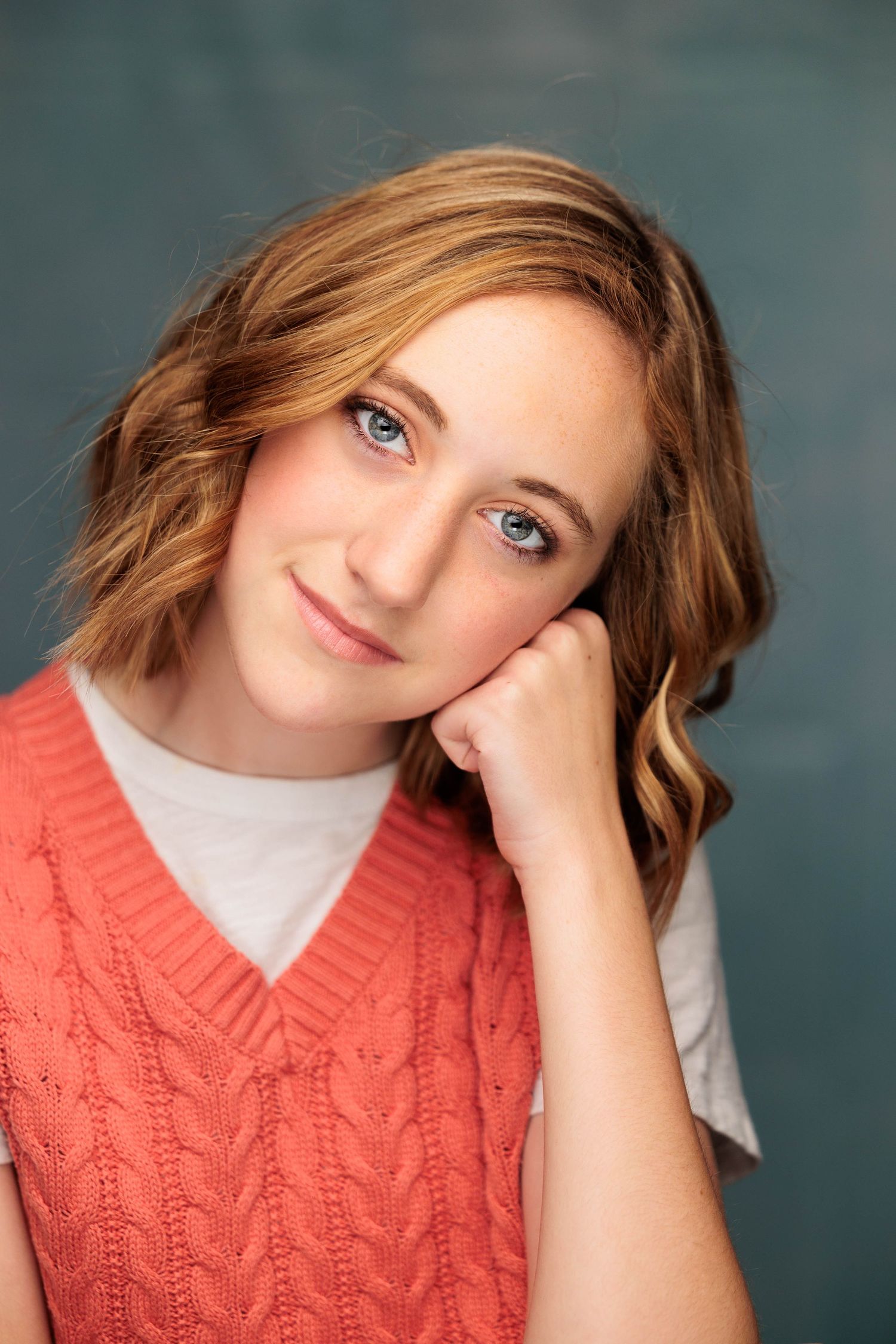 Young girl with blue eyes and auburn hair poses in coral knit vest against gray backdrop.