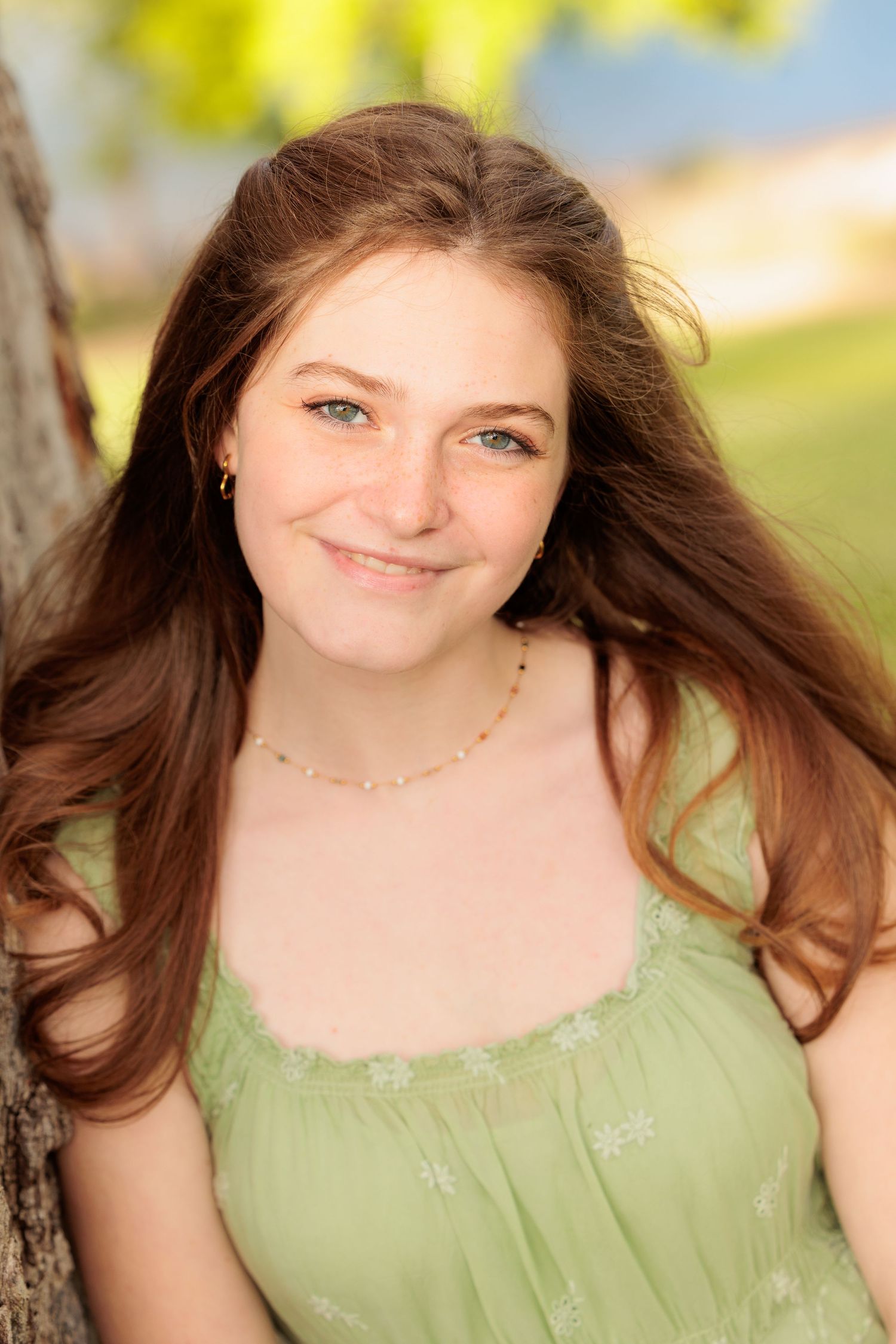 Teenage girl with long auburn hair smiling outdoors in a green dress near a tree.