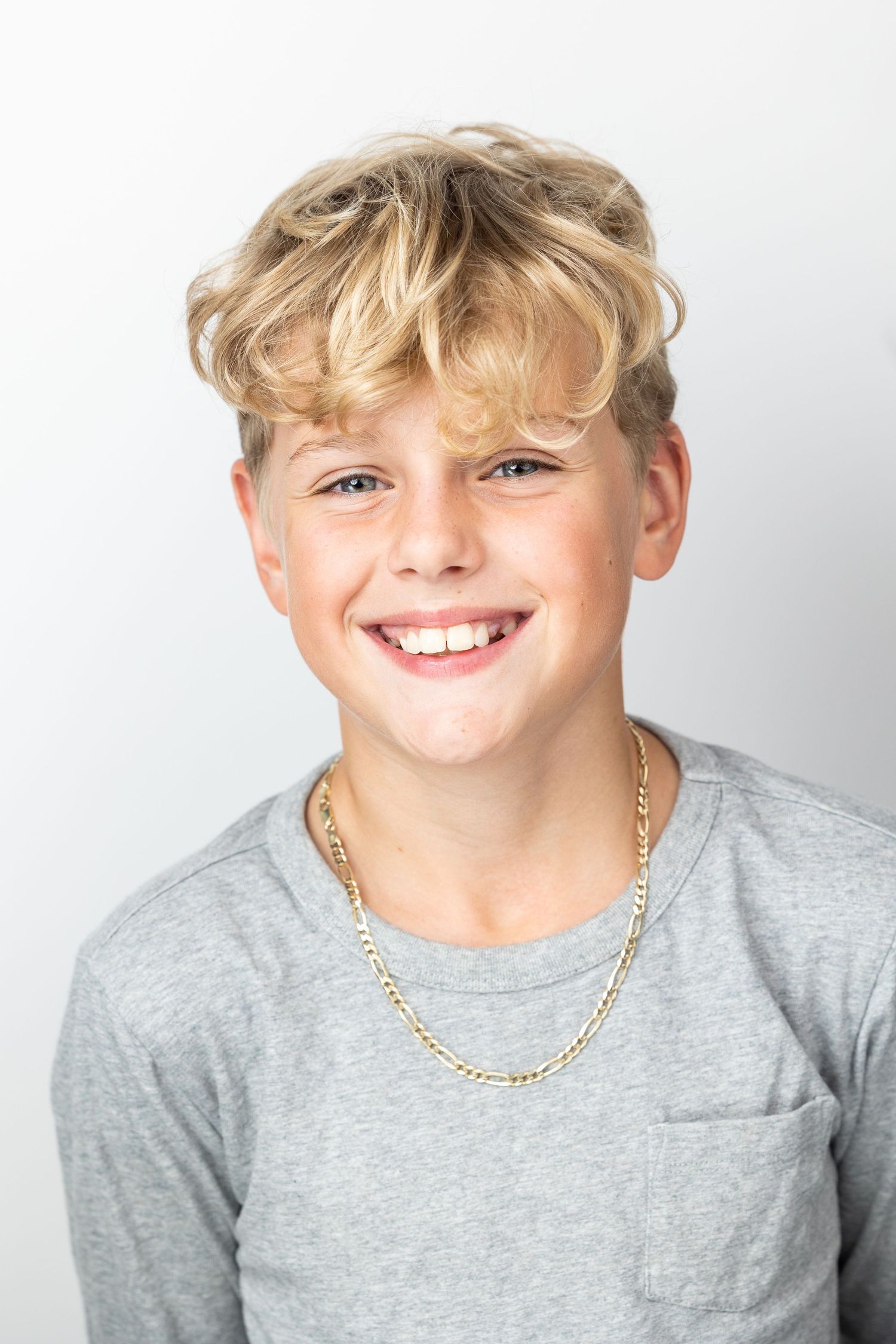 Smiling blond teen boy in gray tee with gold chain poses against white background.