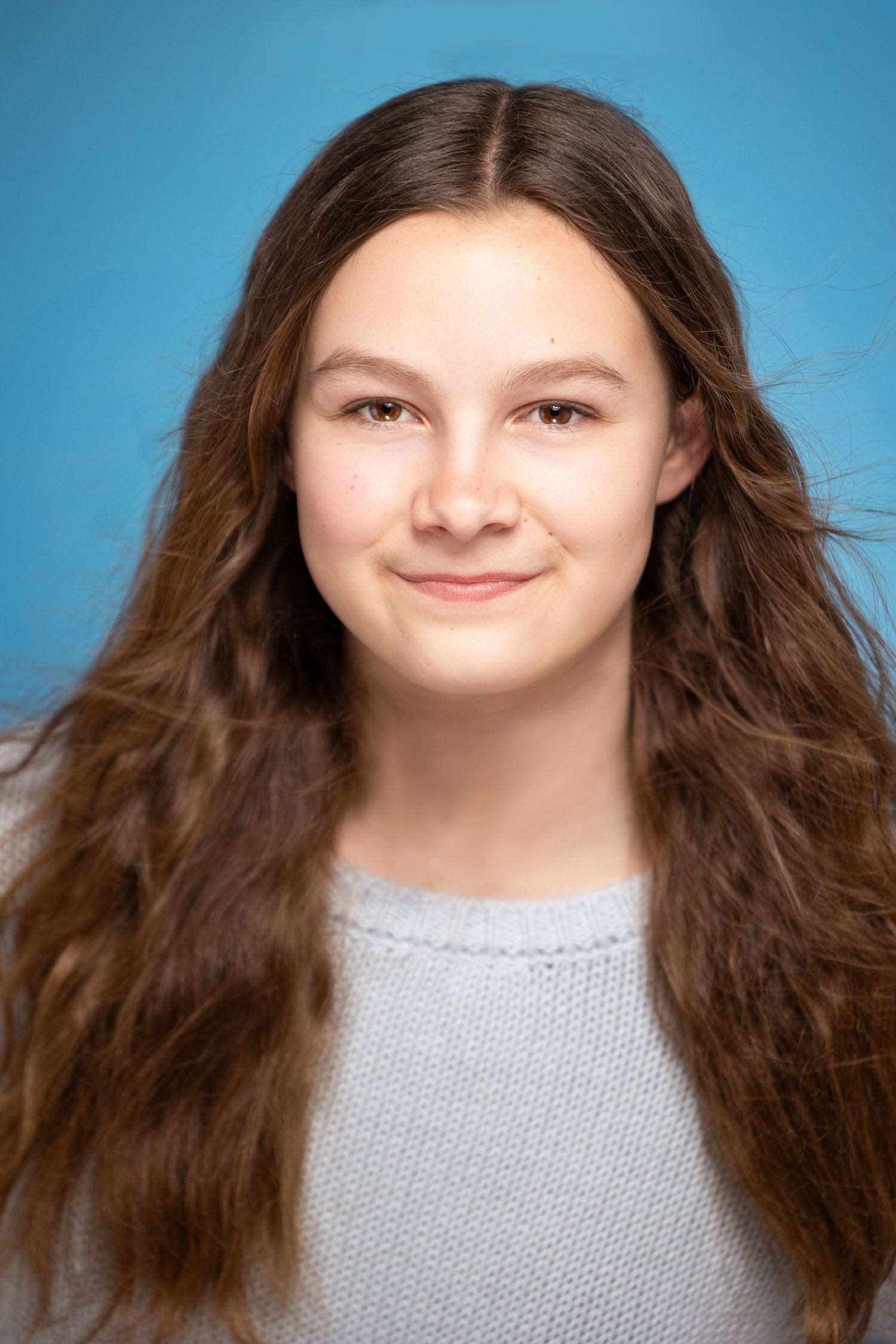 Teen girl with long wavy brown hair smiles softly in gray sweater on blue background.