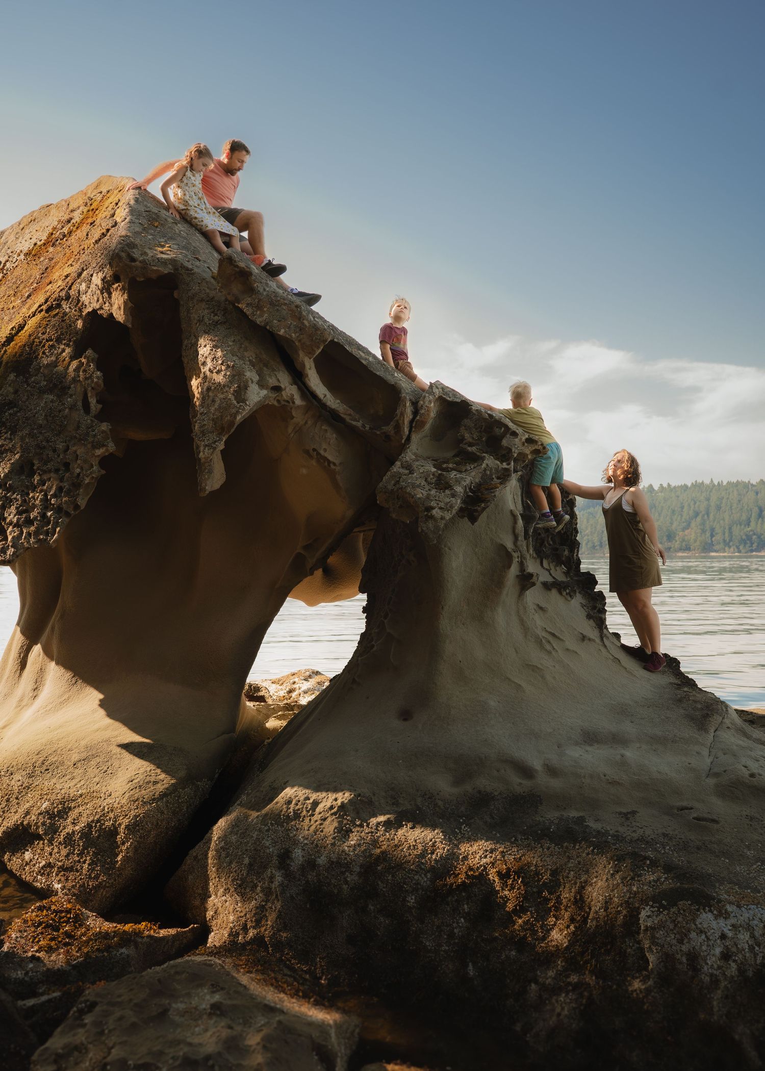Family climbing large weathered sandstone rock formation on a scenic Pacific Northwest beach with ocean and forest backdrop.