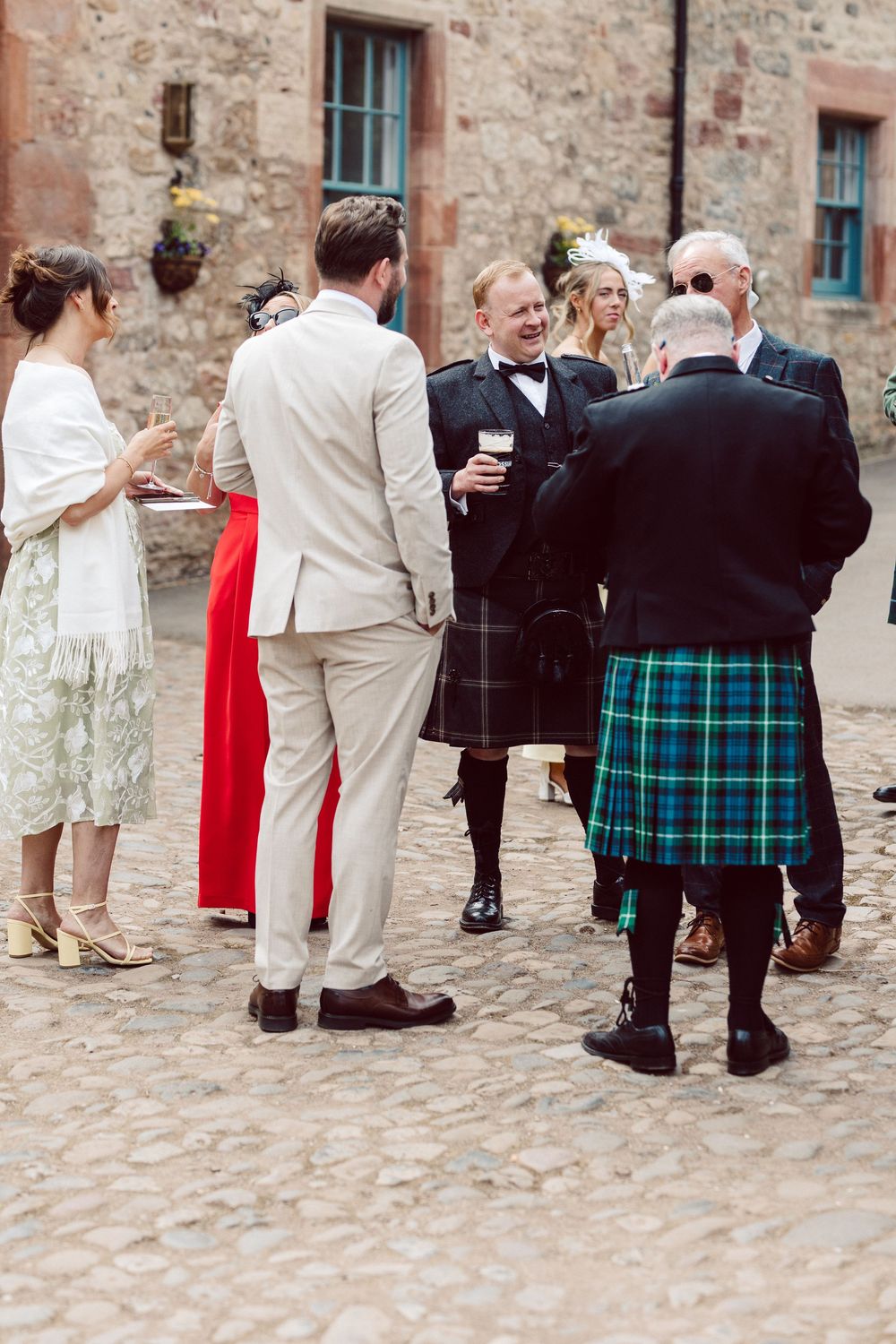 Wedding guests in traditional Scottish kilts and formal attire gather on cobblestone courtyard during reception.