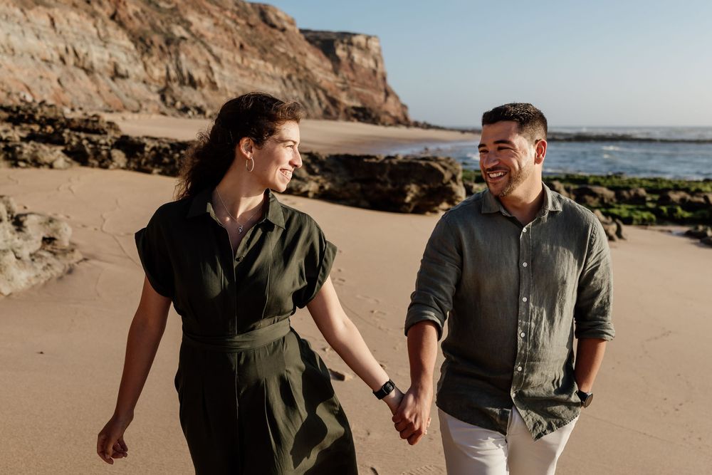 A man and woman walking together on a beach with rocky cliffs in the background.