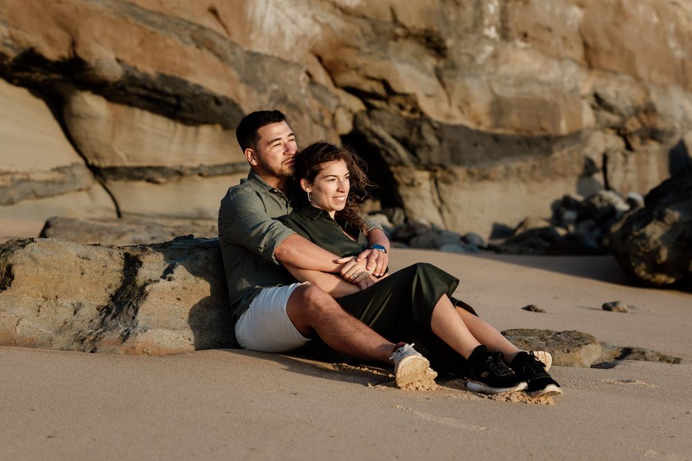 A couple sitting together on a beach with rocky cliffs in the background.