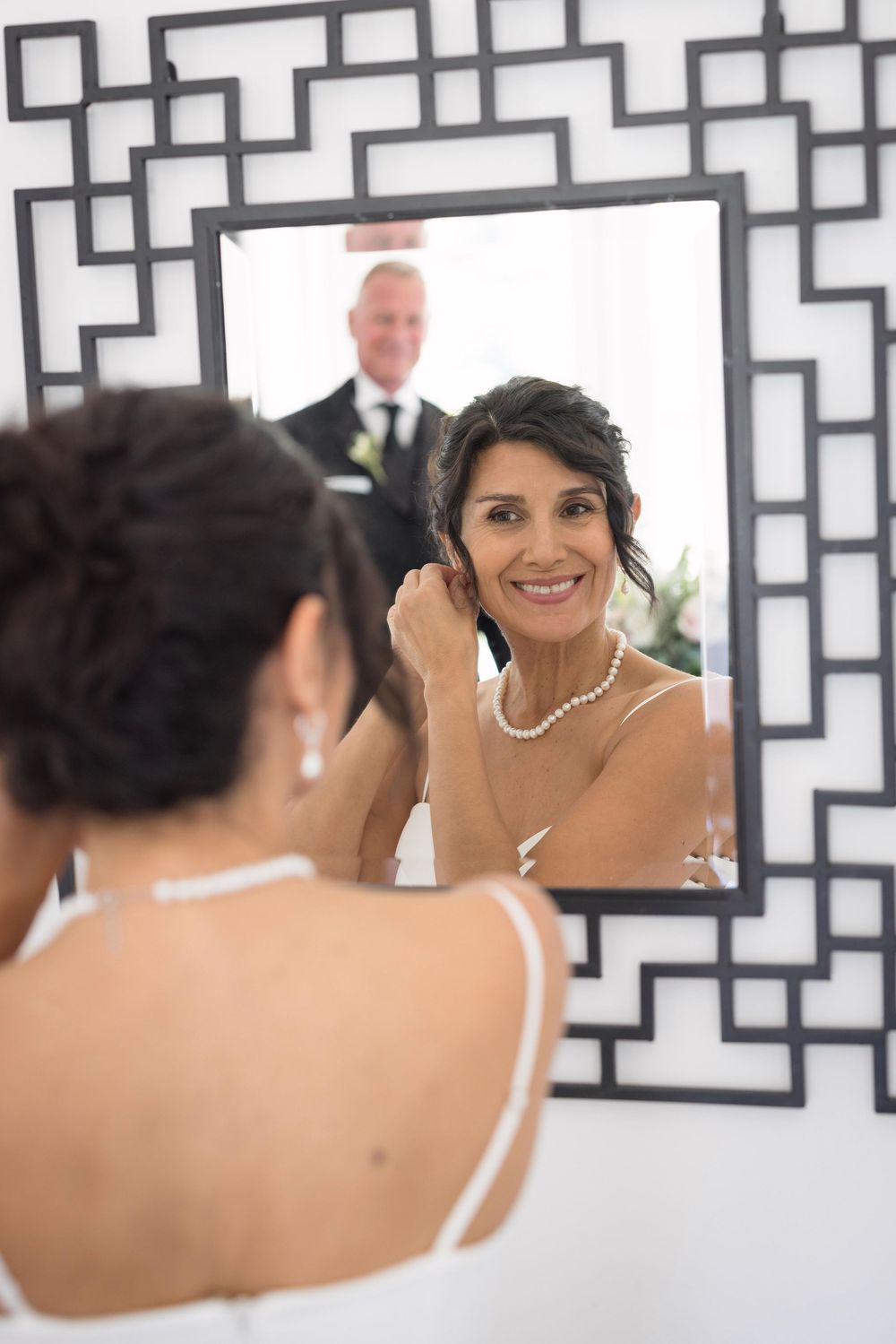 Bride in white dress adjusts pearl necklace while smiling in ornate mirror, groom visible in reflection behind her.