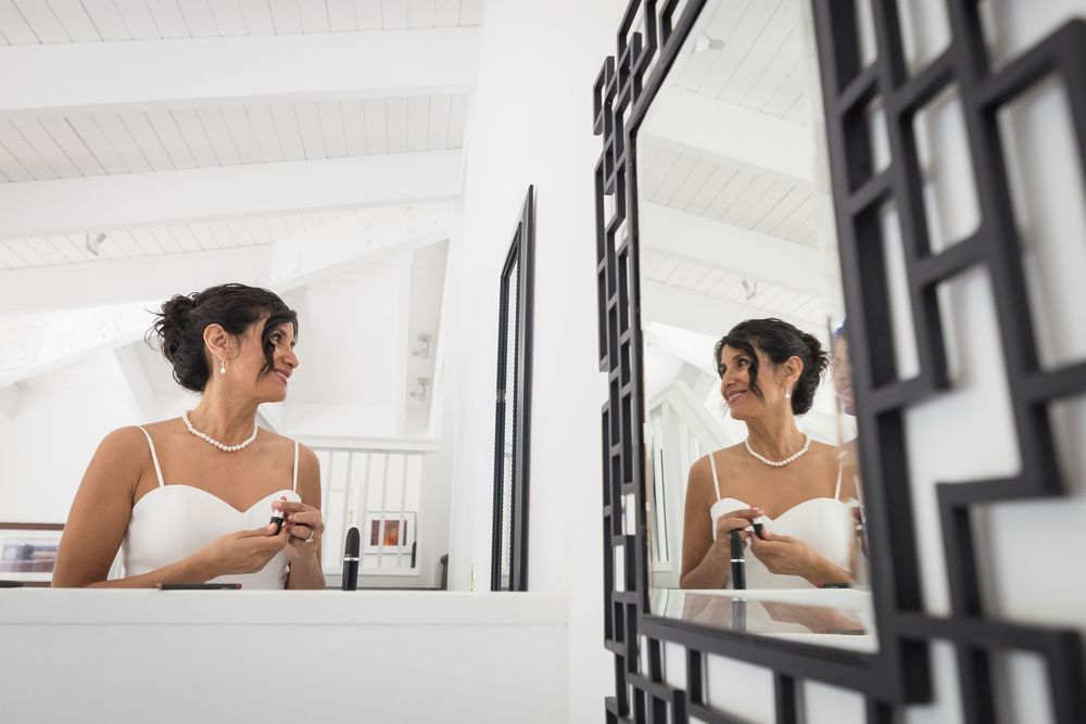 Smiling bride in white dress and pearl necklace applying makeup while her reflection is visible in a decorative mirror.