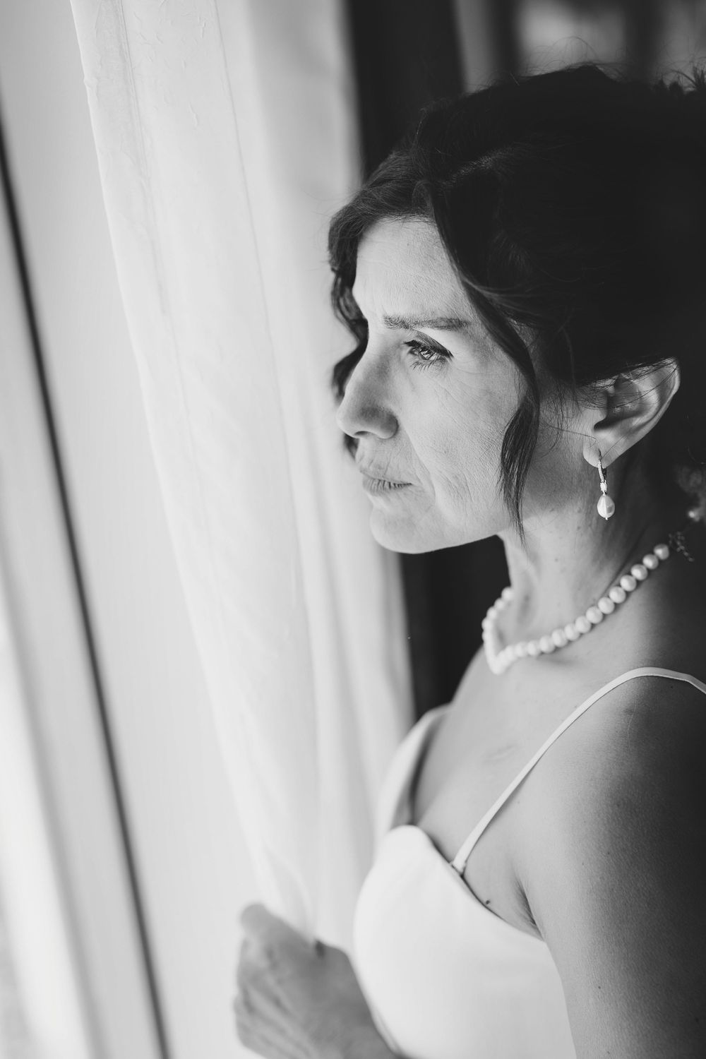 Black and white portrait of a bride in pearl jewelry gazing thoughtfully out a window on her wedding day.