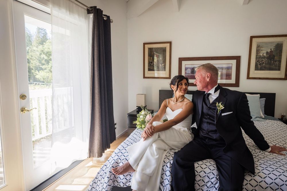 Bride and groom sitting together on a bed, sharing a joyful moment in an elegantly decorated room.