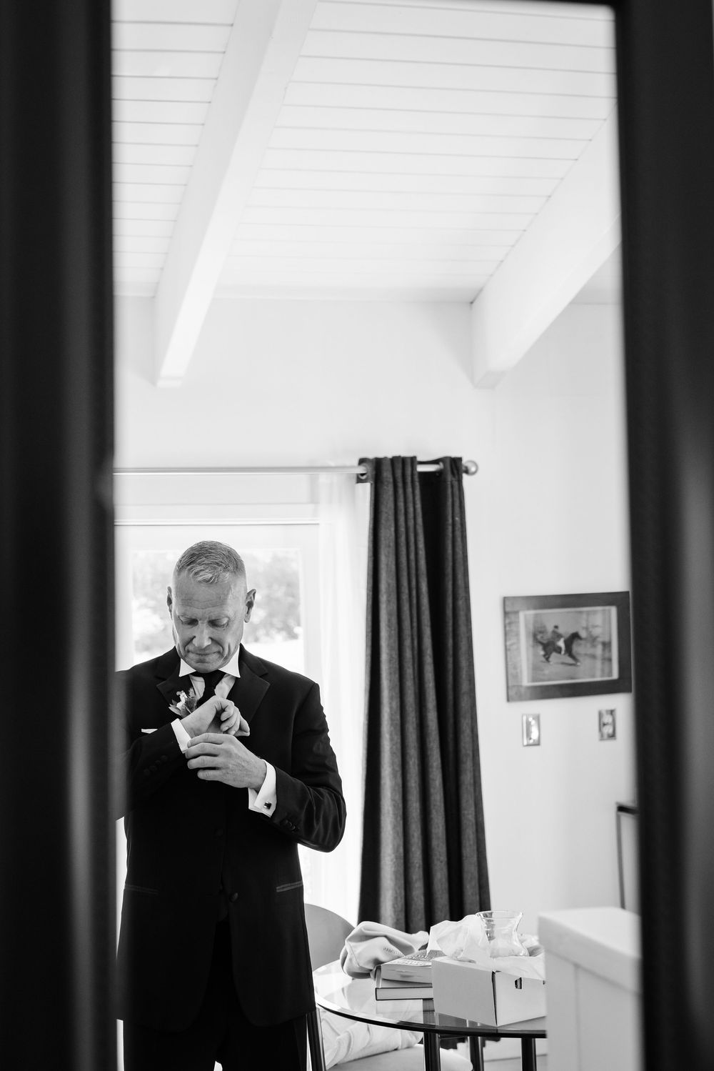 Black and white photo of a man in a suit adjusting his cufflinks in a bright room.