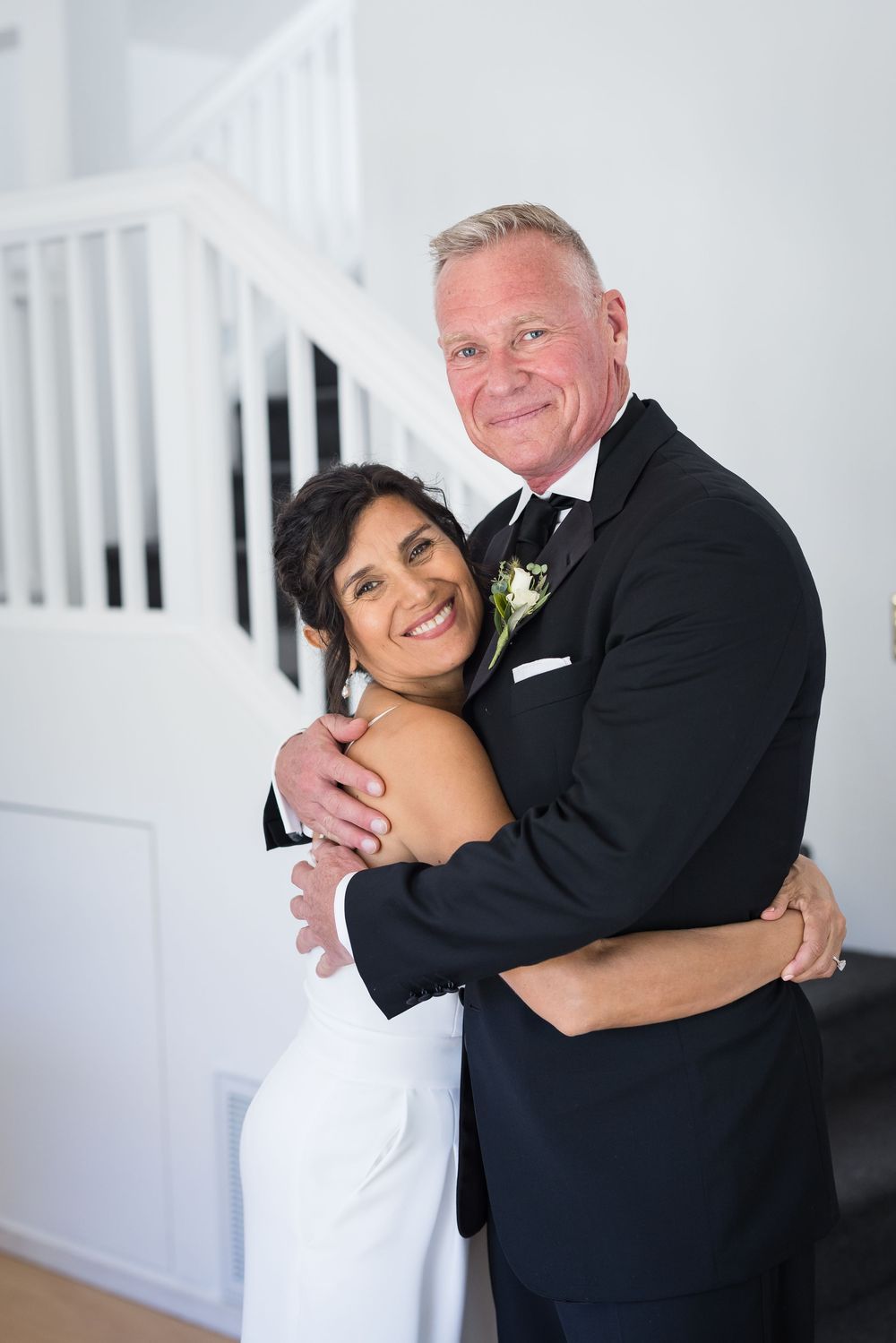 Smiling bride and groom embrace indoors on their wedding day, groom in black tuxedo, bride in white gown.