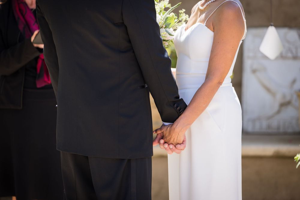 Bride and groom holding hands during outdoor wedding ceremony, wearing white dress and black suit.