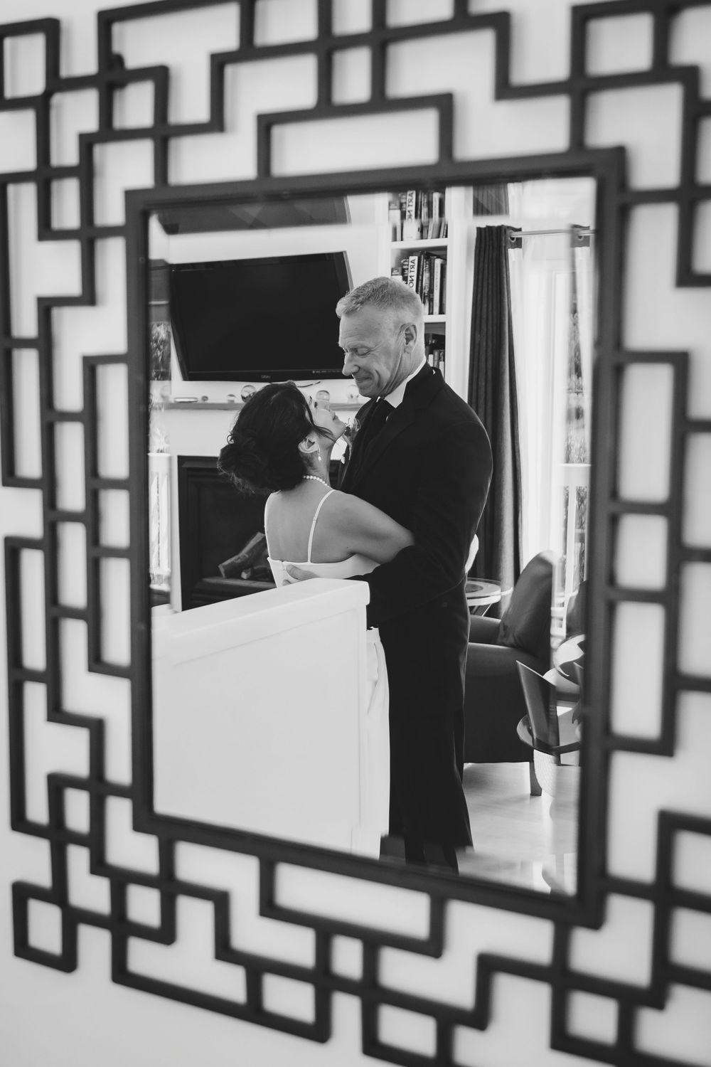 Black and white mirror reflection of a couple in wedding attire sharing a romantic embrace indoors.