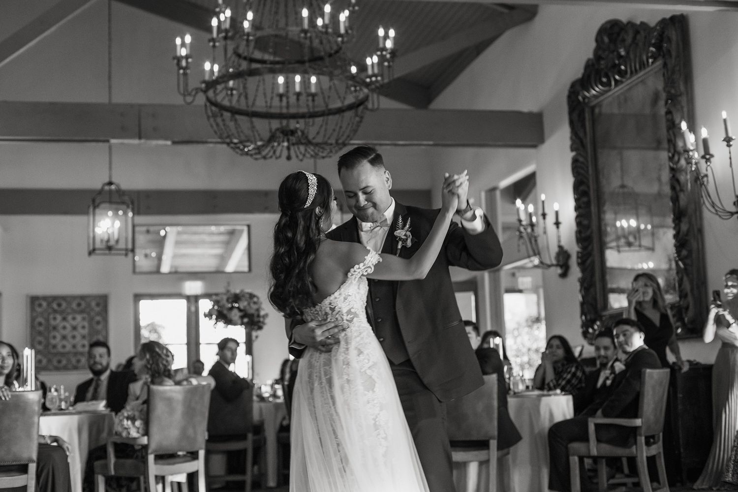 Black and white photo of a couple sharing their first dance at a wedding reception with guests seated around them.