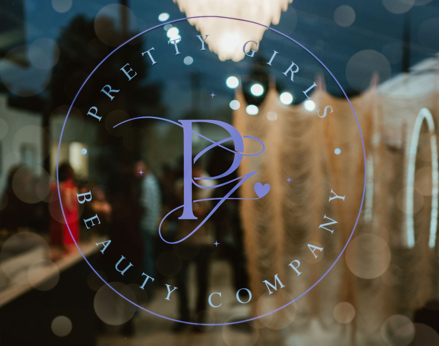 Pretty Girls Beauty Company logo etched on a glass storefront window with bokeh background.