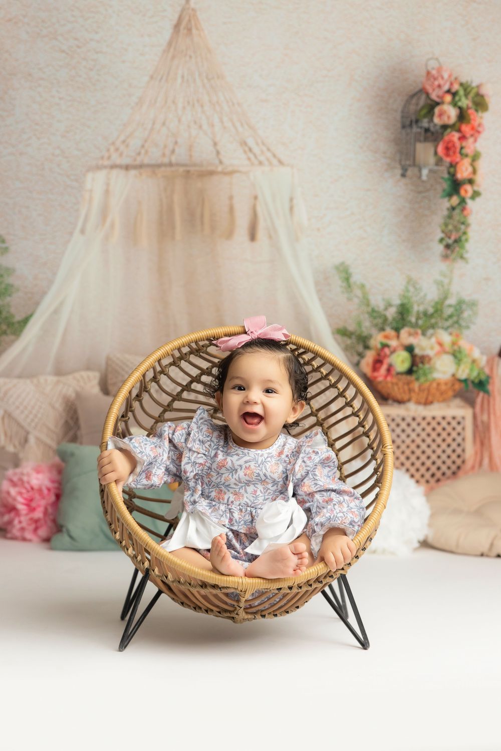 Smiling baby girl in floral outfit sitting in round rattan chair with boho nursery decor backdrop.