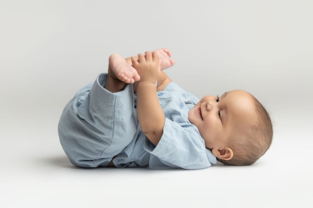 A cute baby in a blue onesie lying on its back, happily playing with its feet against a white background.
